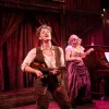 Two performers play ukuleles on stage for Calamity Jane, set against rich red drapes and wooden paneling. One sings passionately, while the other smiles, creating a lively, theatrical atmosphere.
