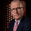 A middle-aged white man wears round glasses and suit with tie. He sits in front of an organ.