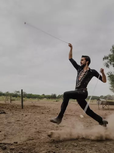 A person in a black outfit holding a whip, mid stride in a dusty paddock.