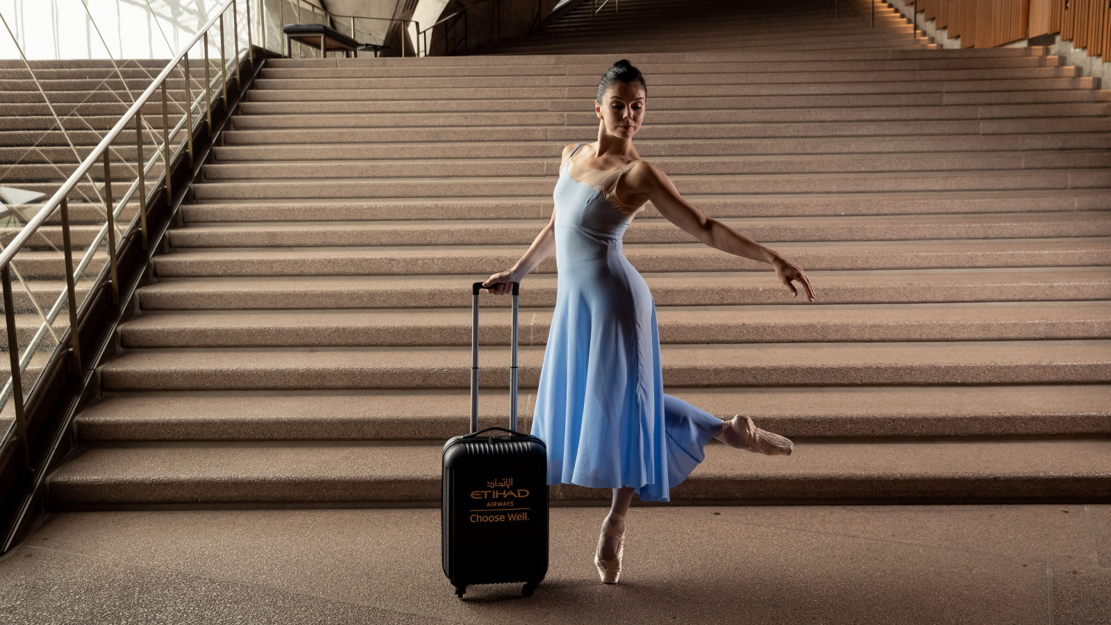A ballerina in a blue dress posed with a suitcase on the steps of the Sydney Opera House.