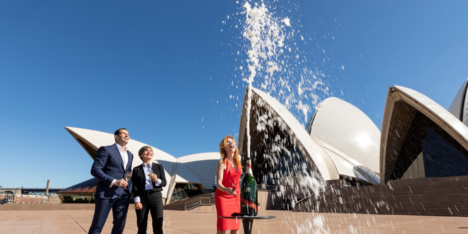 A woman in a red dress and two people in suits standing outside the Sydney opera House, watching a bottle of Champaigne spray into the air.