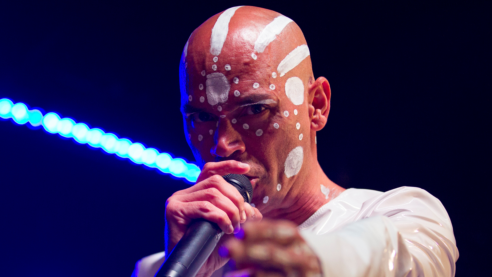 A bald man with aboriginal face makeup singing into the mic.