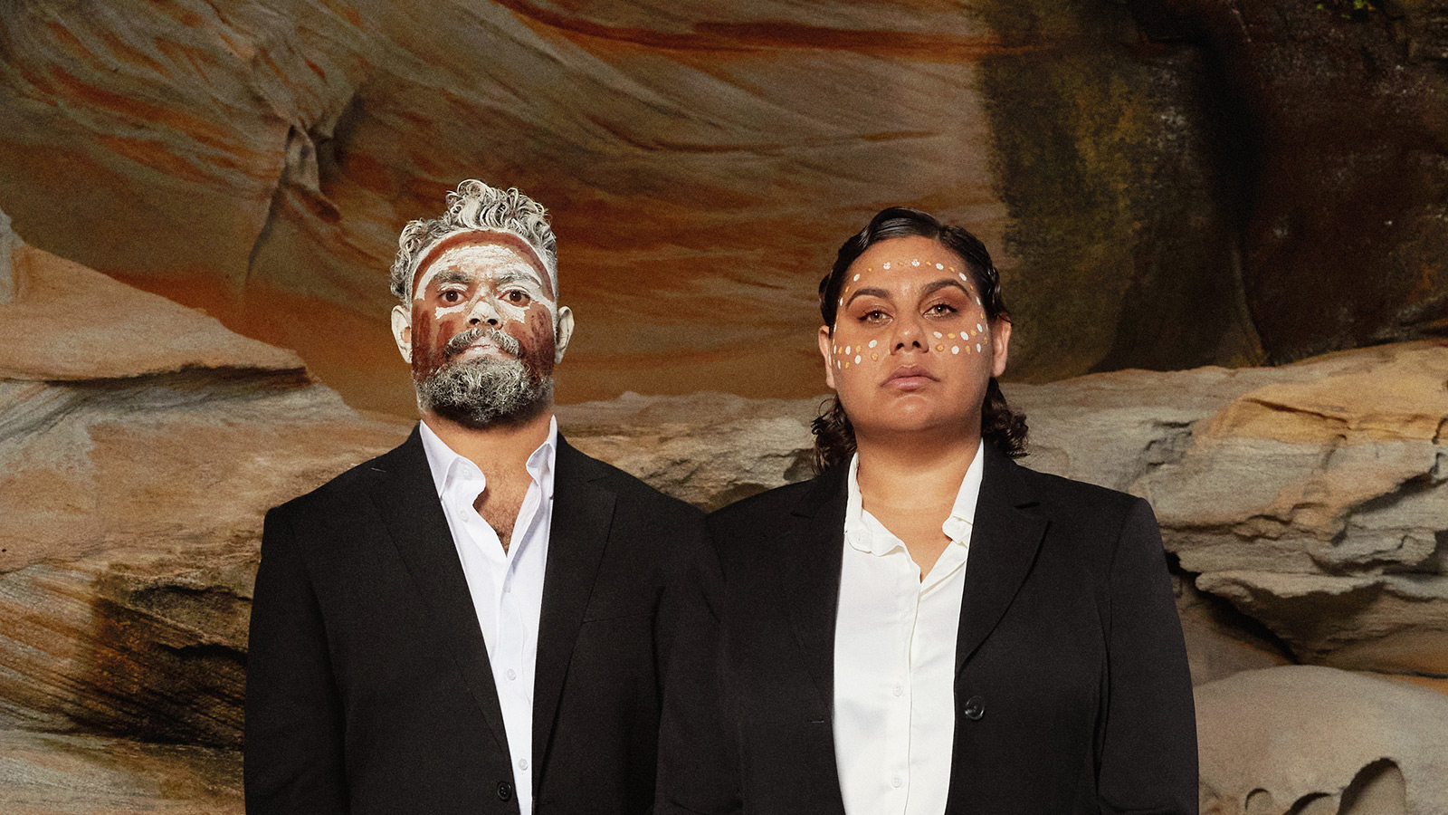 An aboriginal man and women wearing a black suit with traditional face art.