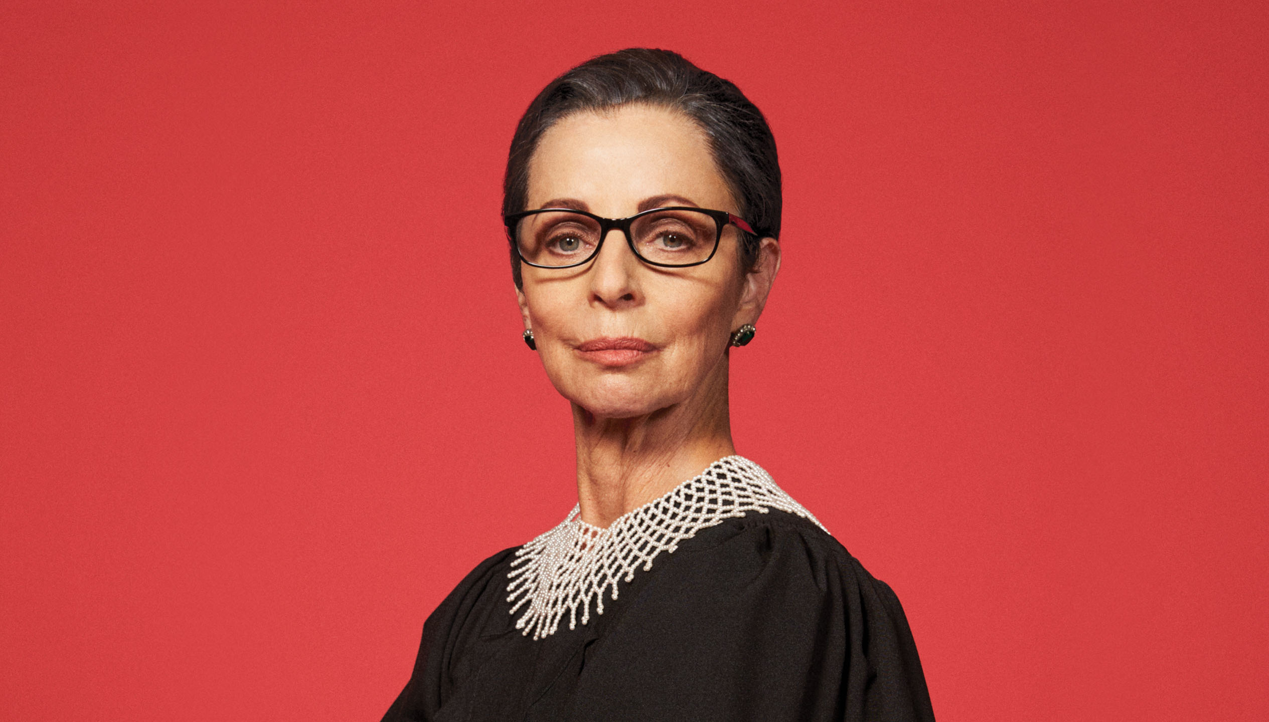 A white woman in her 60s has her hair sleekly pulled back and pursed lips wears black glasses and a lawyer's black robe with white crocheted neck piece. She stands in front of an all-red background.