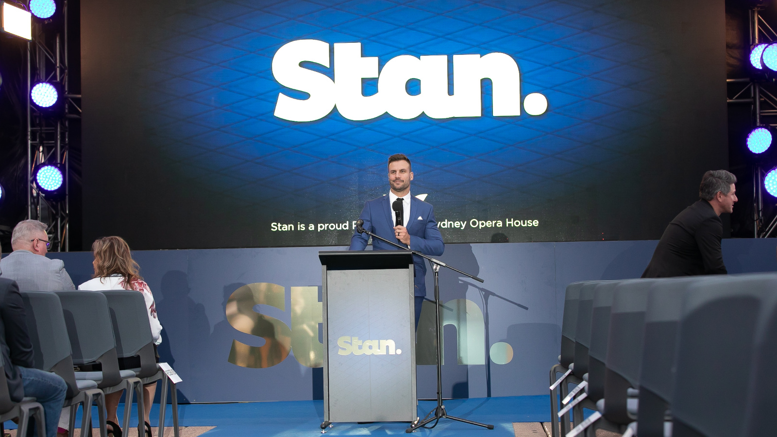 A man in a blue suit standing behind a speaking podium on a stage.