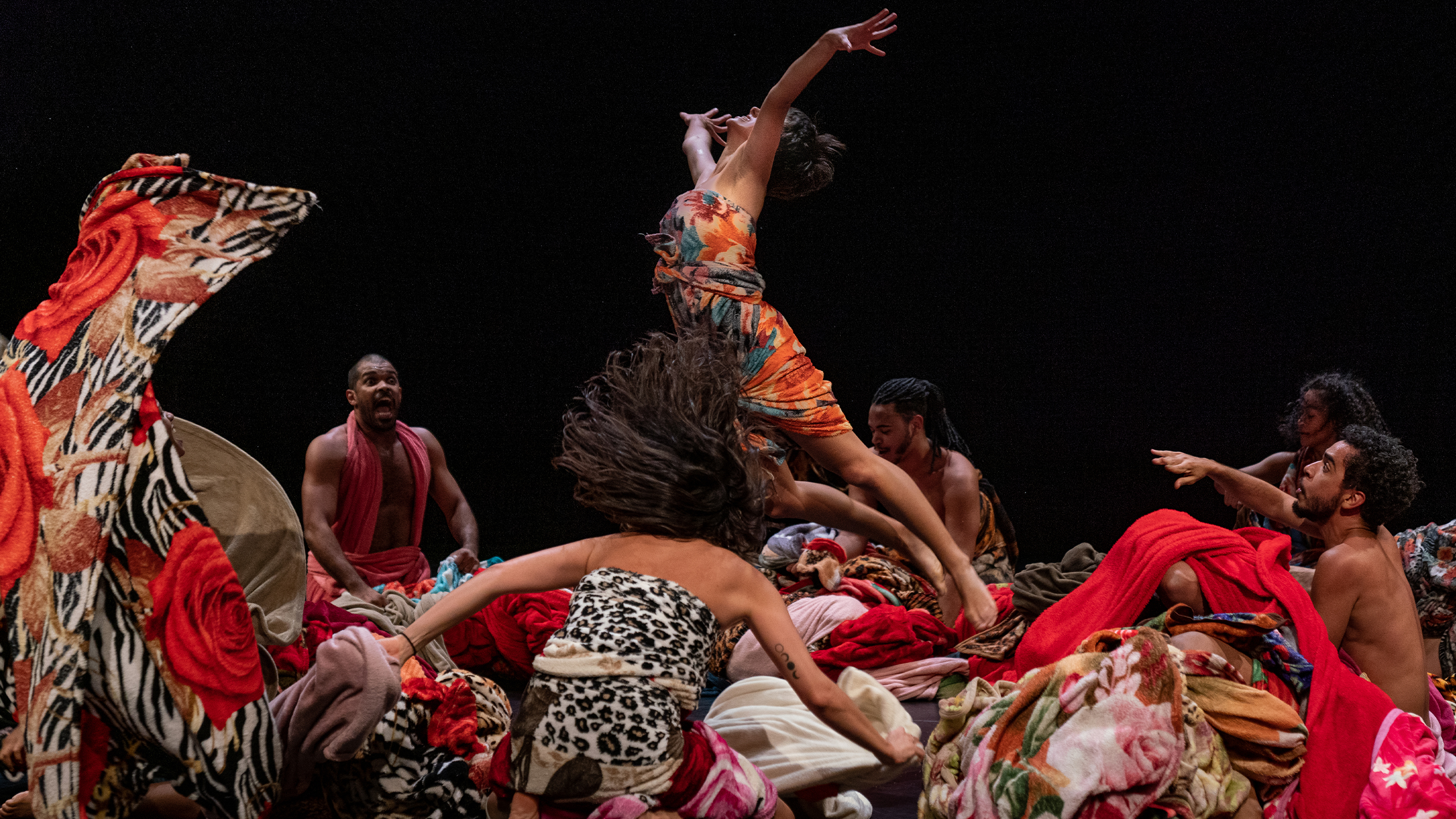 A group of Brazilian dancers, men and women, wear clothes with red and animal print patterns. They are on stage and a female dancer is mid-air as if just thrown by the male dancers.