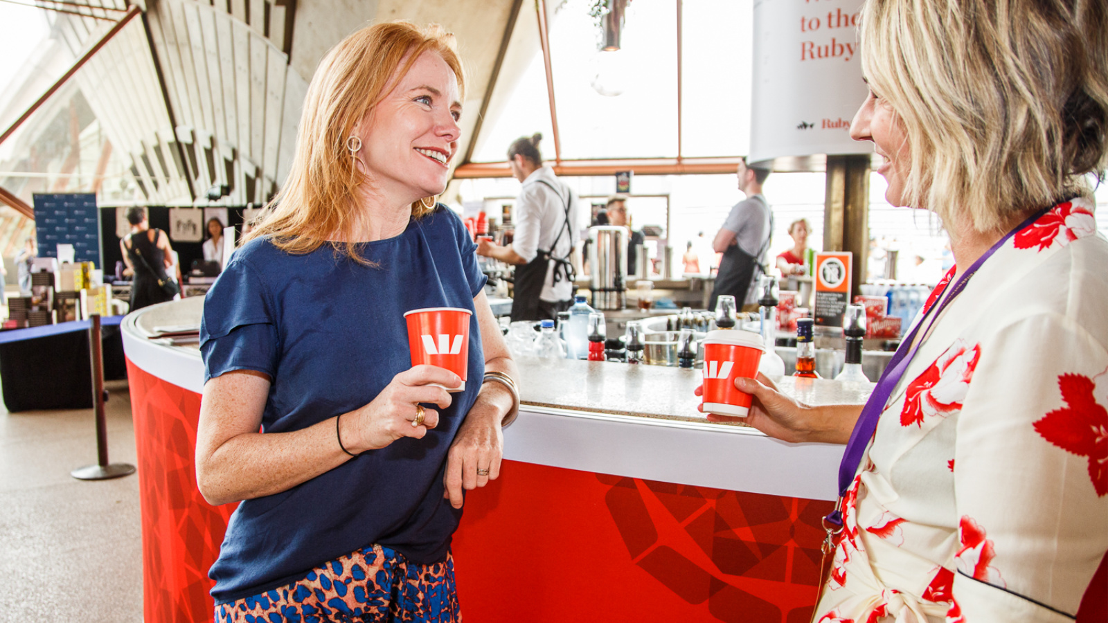 Two women holding coffee cups and talking in front of a bar.