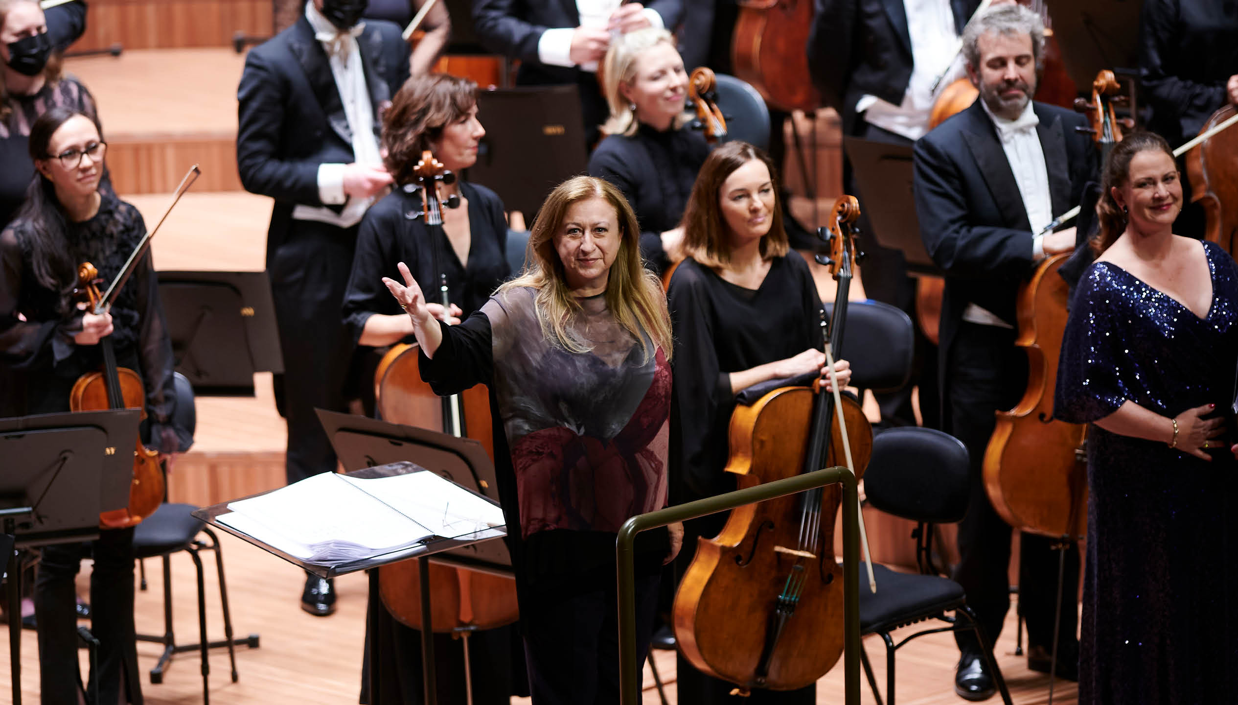 A woman in her 60s with long brown hair stands in front of an orchestra at the conductors desk, waving to the orchestra.