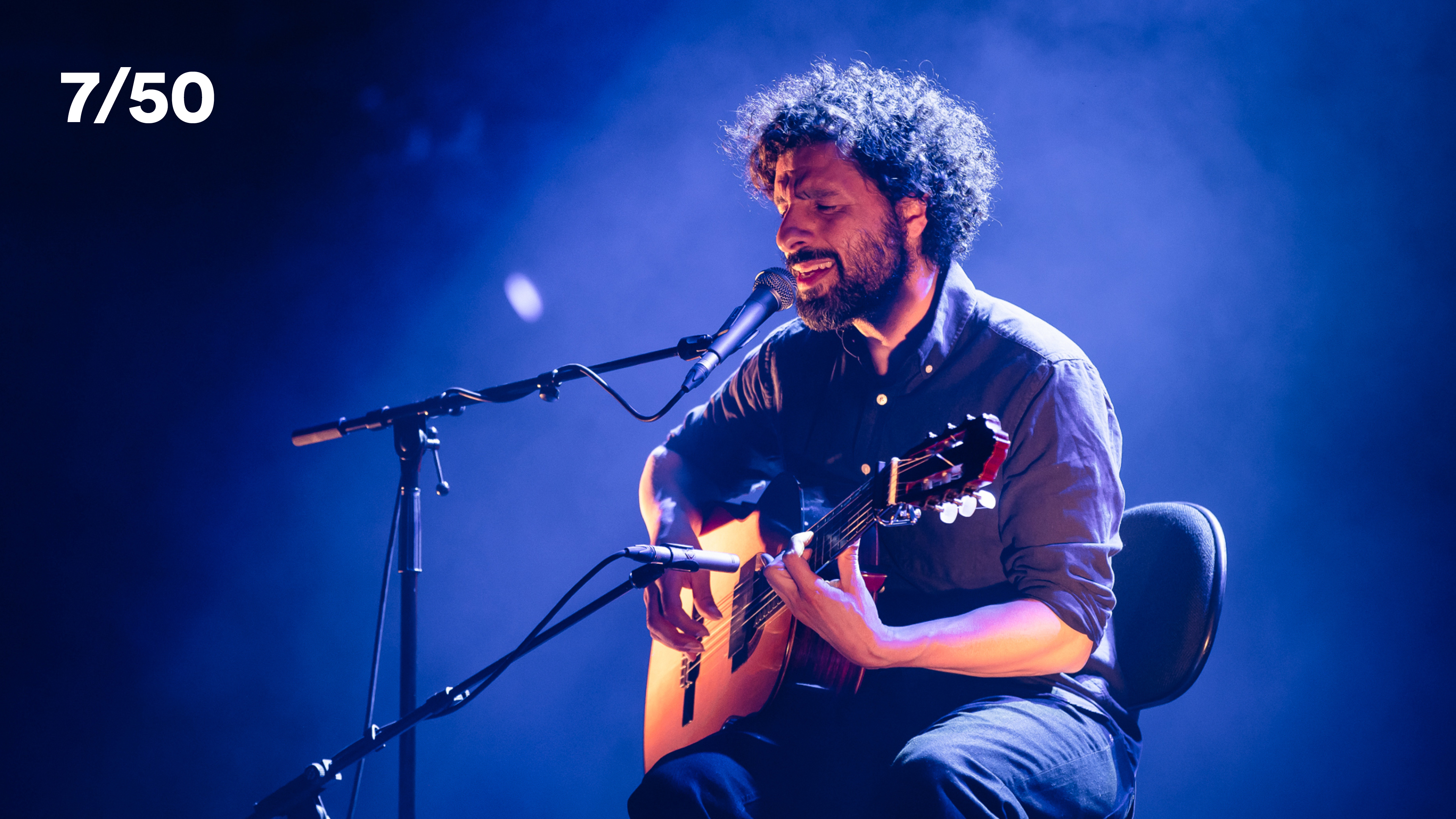 A signer performing on stage under blue lighting, sitting on a chair signing and playing guitar.