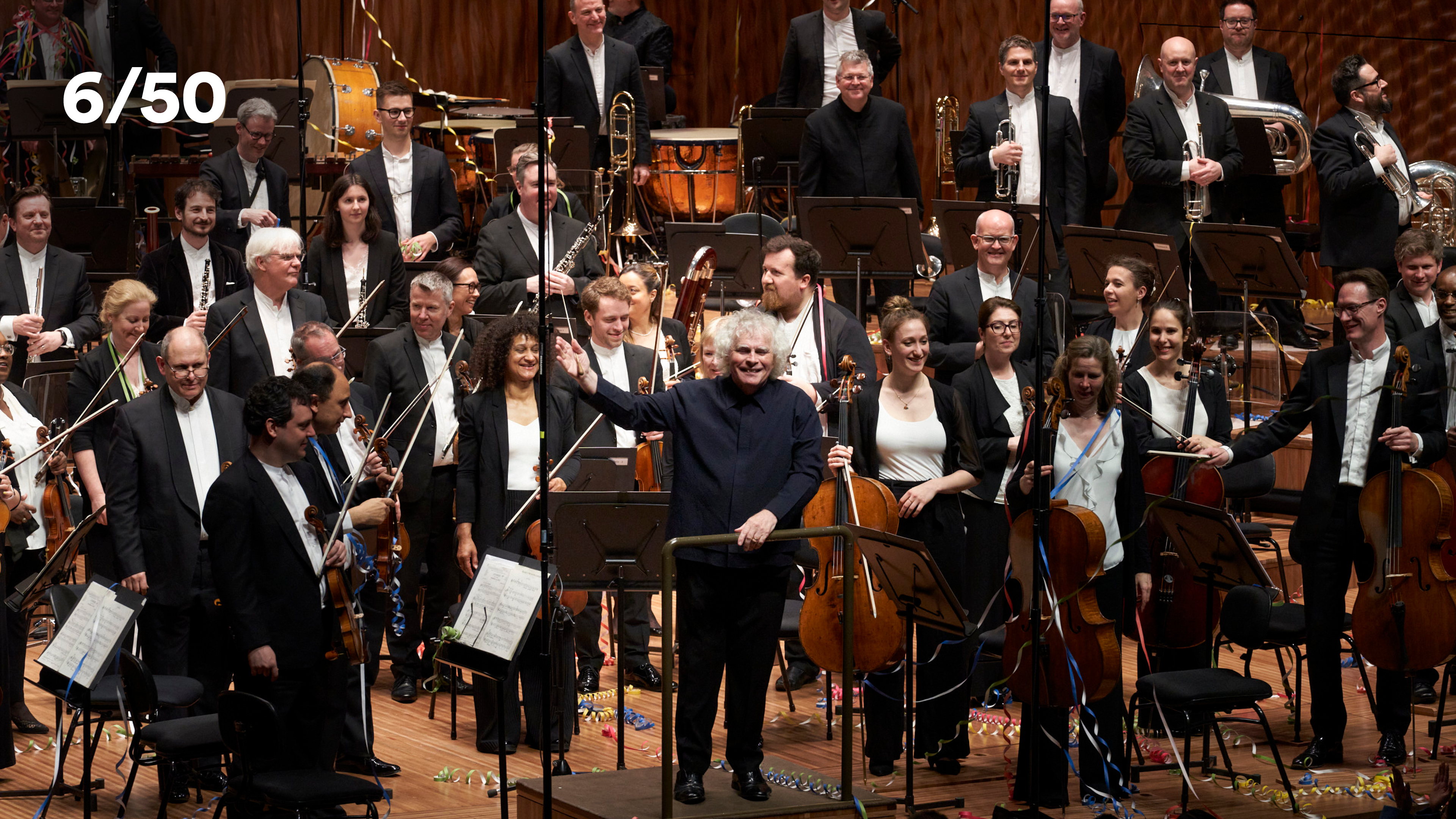 An orchestra on stage, members of the orchestra are standing, the conductor is waving their right arm, smiling.