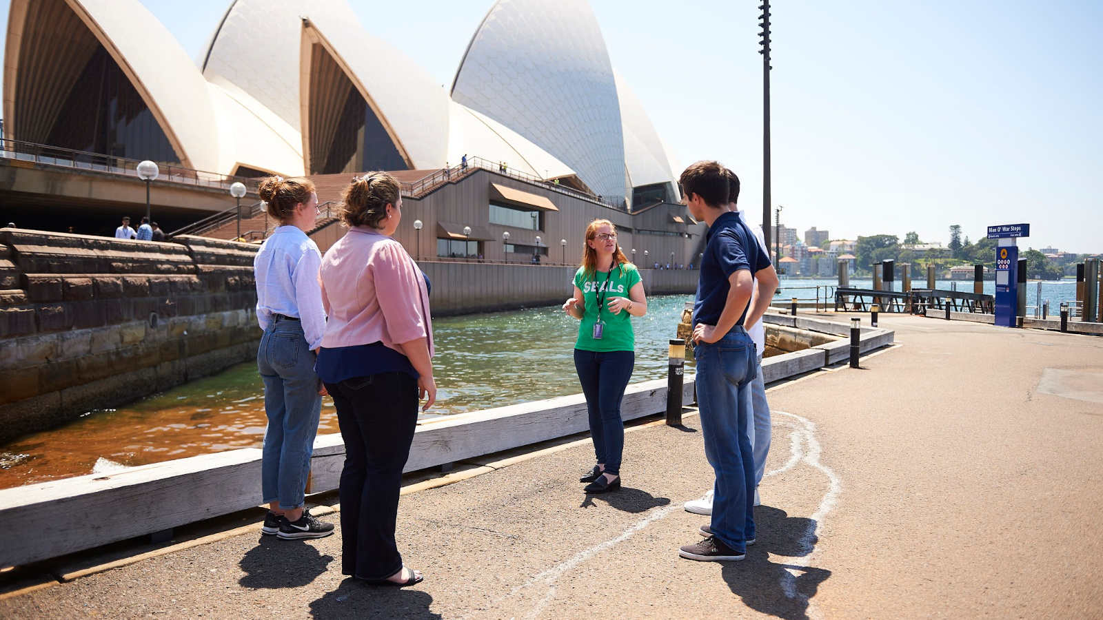 A woman standing outside with four other people, giving a tour of the Sydney Opera House.