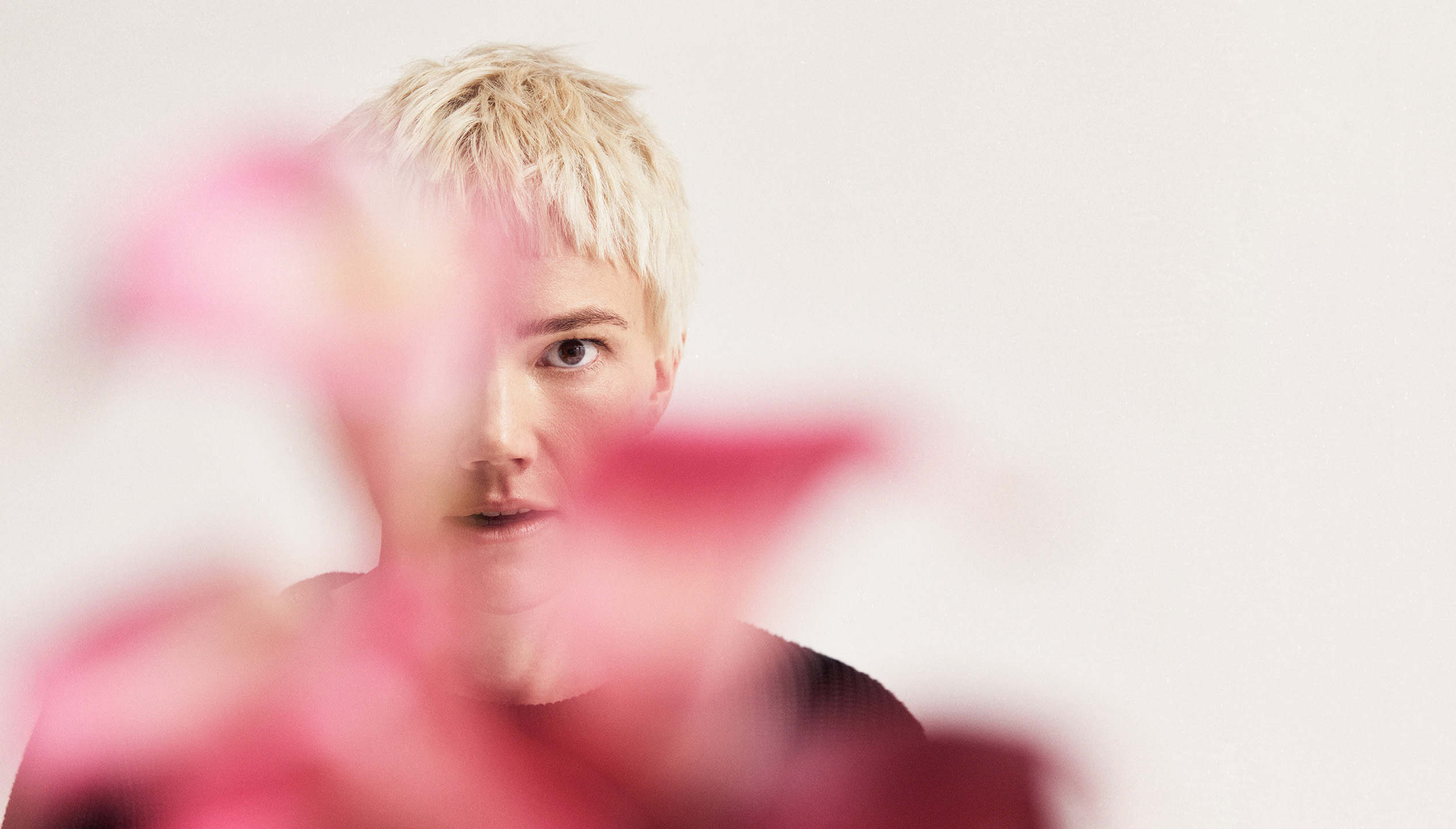 A person with short blonde hair standing behind some pink foliage against a white background.