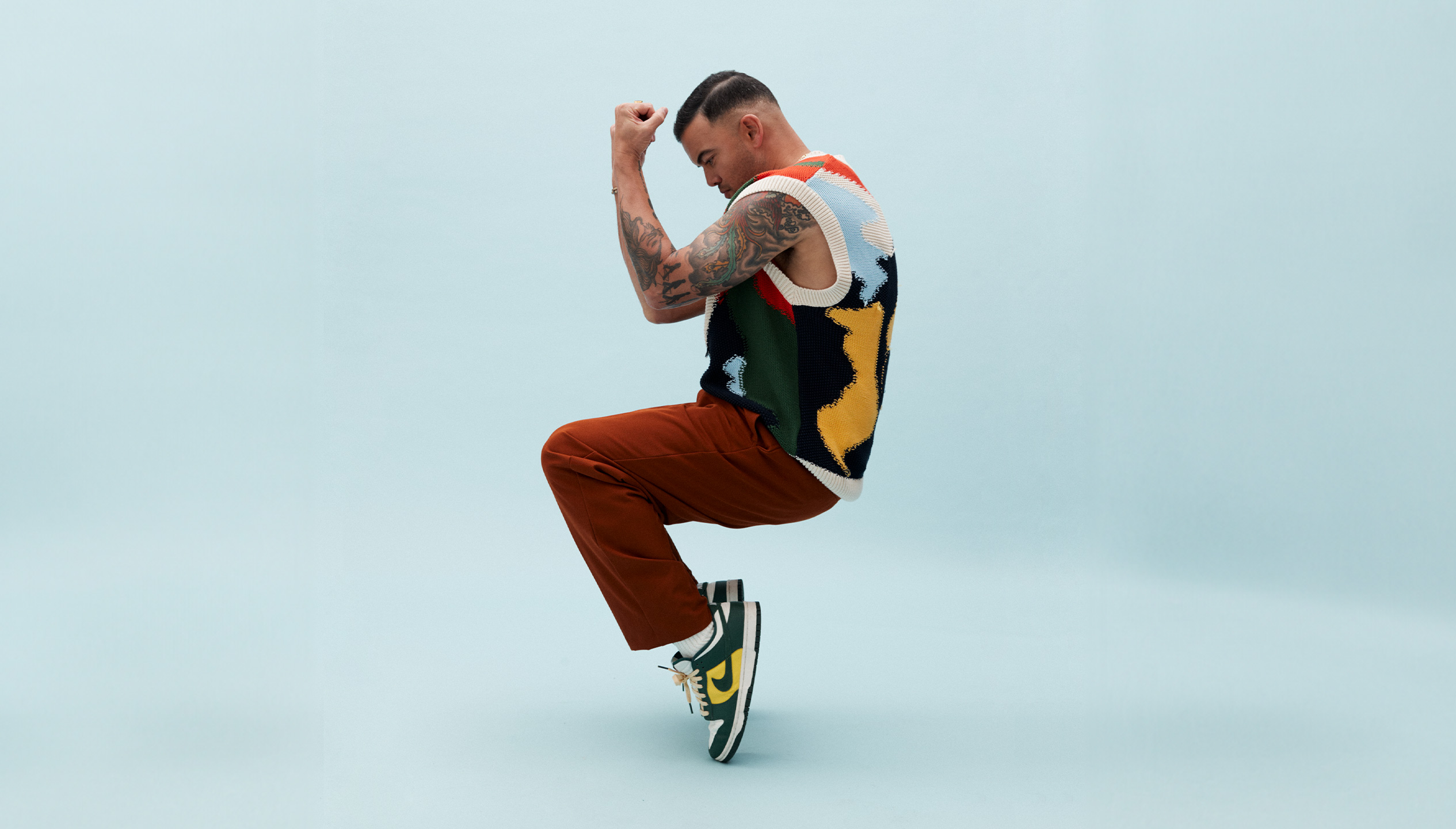 A man with tanned skin and short black hair is posed standing on his toes, with bent knees and his arms held up in front of his face. He wears maroon pants, green and yellow Nike shows and a multi coloured vest.