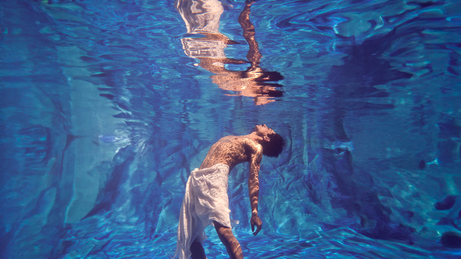 A shirtless man swimming underwater in a pool wearing a cloth skirt has his face looking up towards the sky,