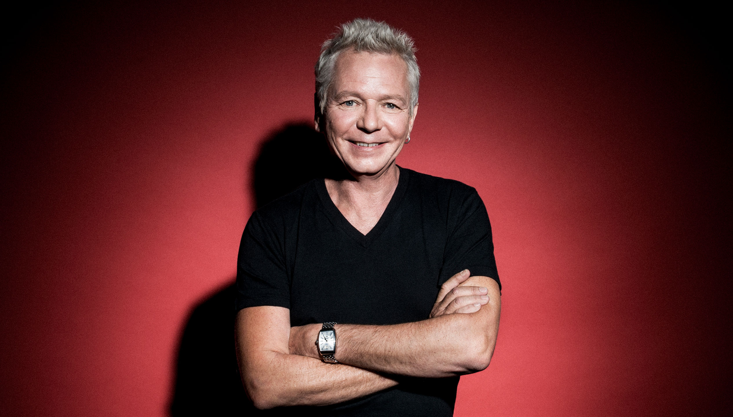 A white man in his 60s with grey hair wears a black t-shirt and has his arms crossed, standing in front of a red background.
