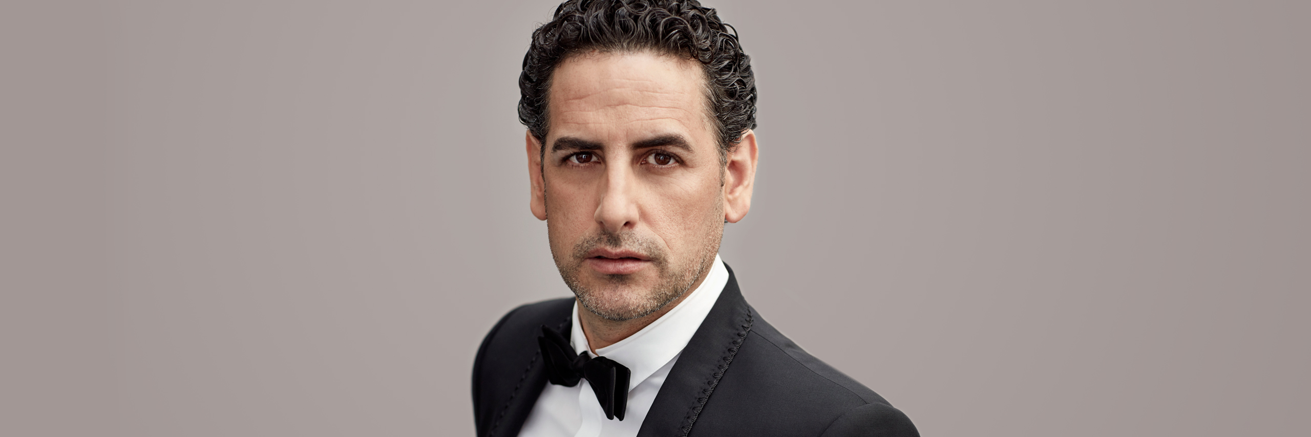A man with dark curly hair looks at the camera wearing a tuxedo