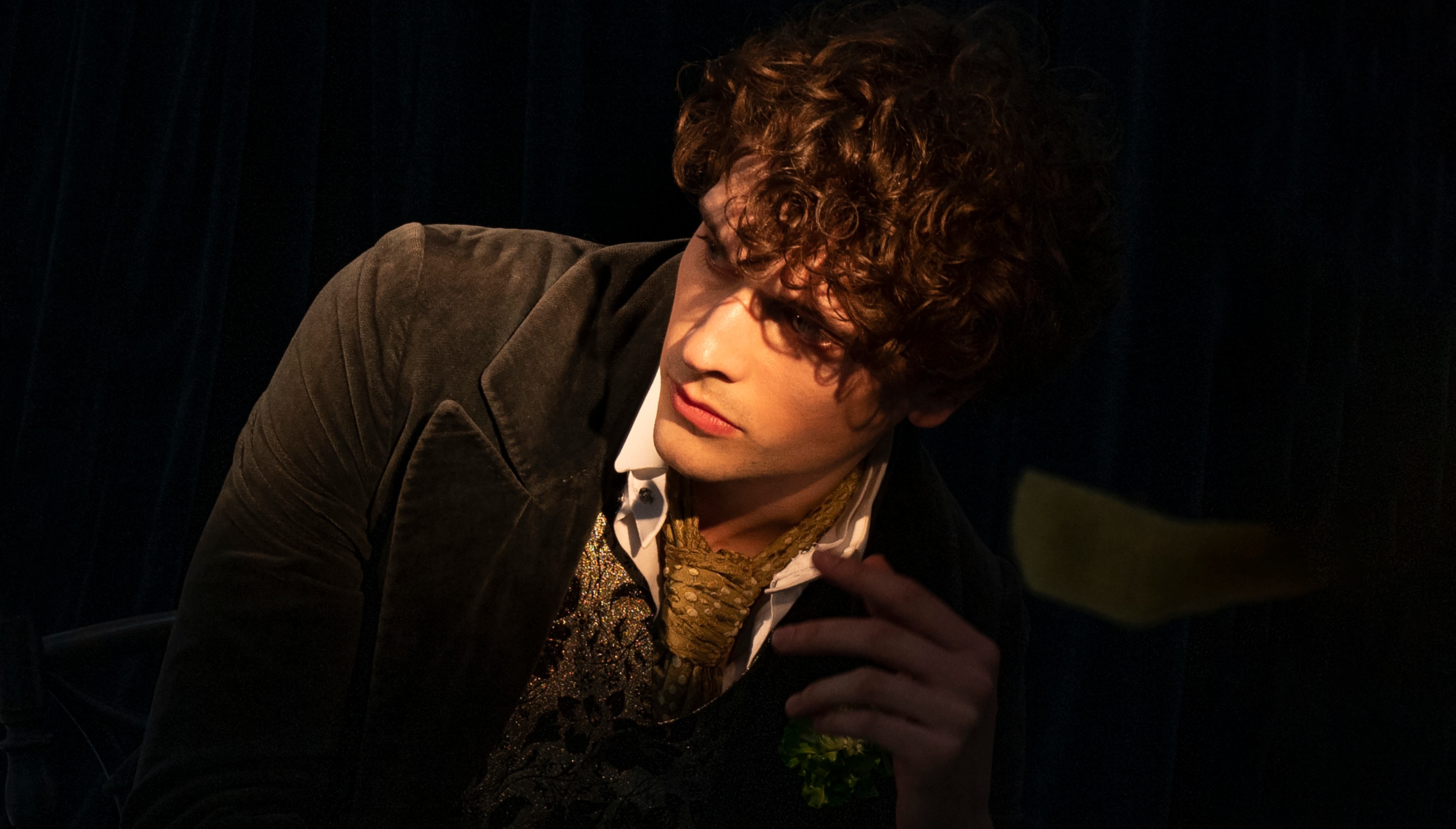A man with light brown curly hair looks off into the distance in front of a black background. He wears a gold tie, white shirt and green jacket.