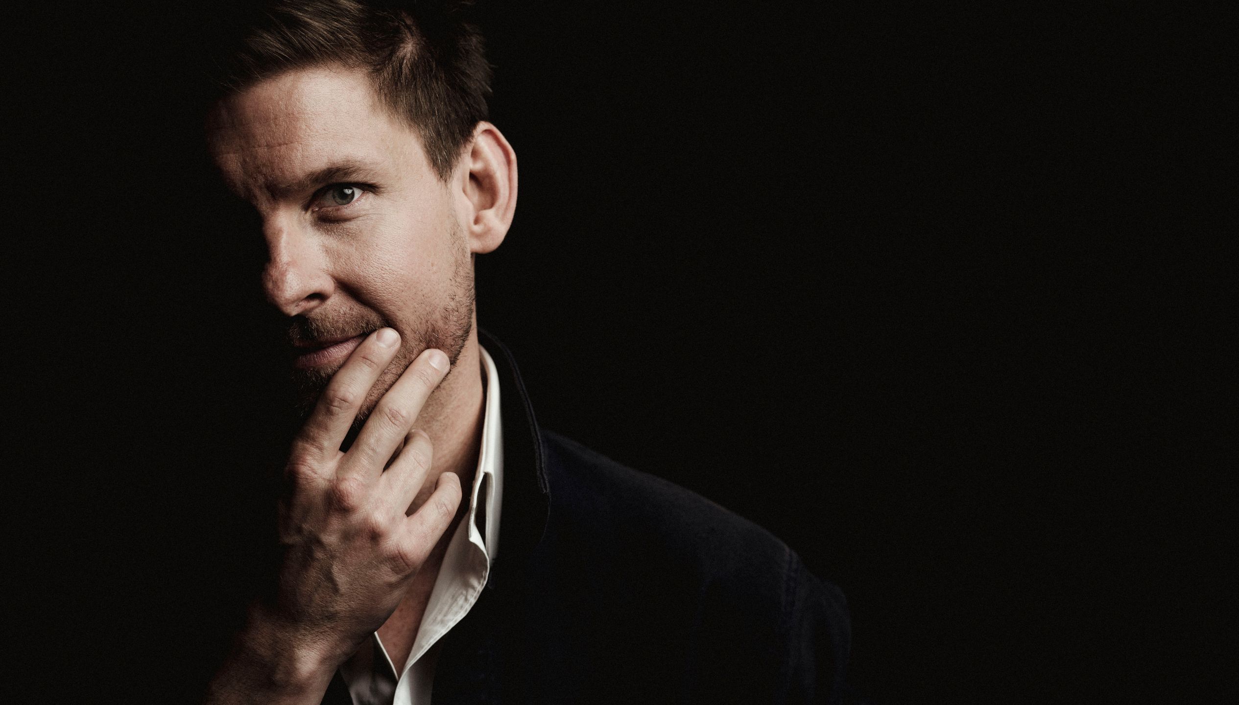 A close up of a white 30-40 year old man wearing a suit has his hand on his chin. Half his face is in darkness.