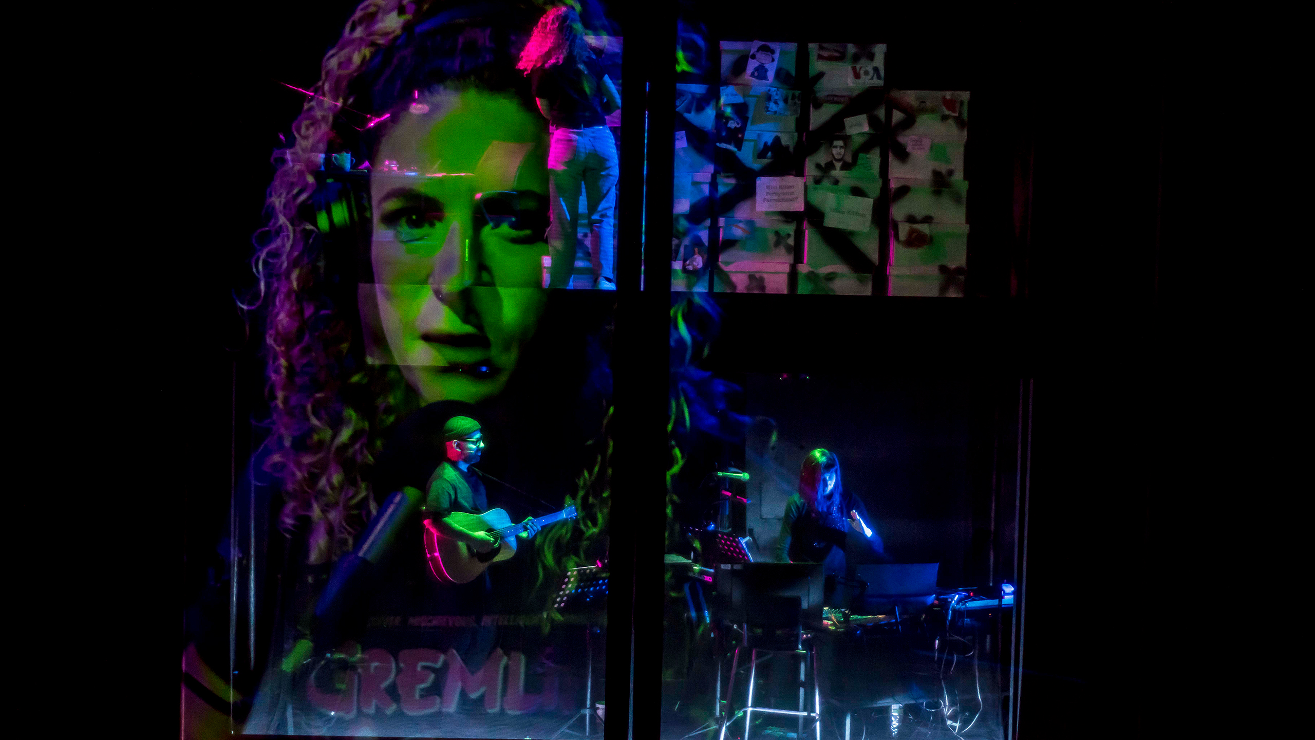 A man wearing a beanie plays the guitar while a woman next to him plays a keyboard, They are surrounded by a projection of a woman's face, and the lighting is green, purple and blue.