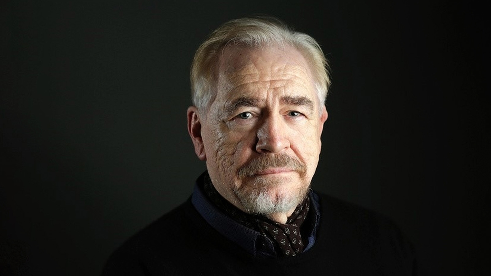 An old man with grey hairs and beard wearing a black shirt.