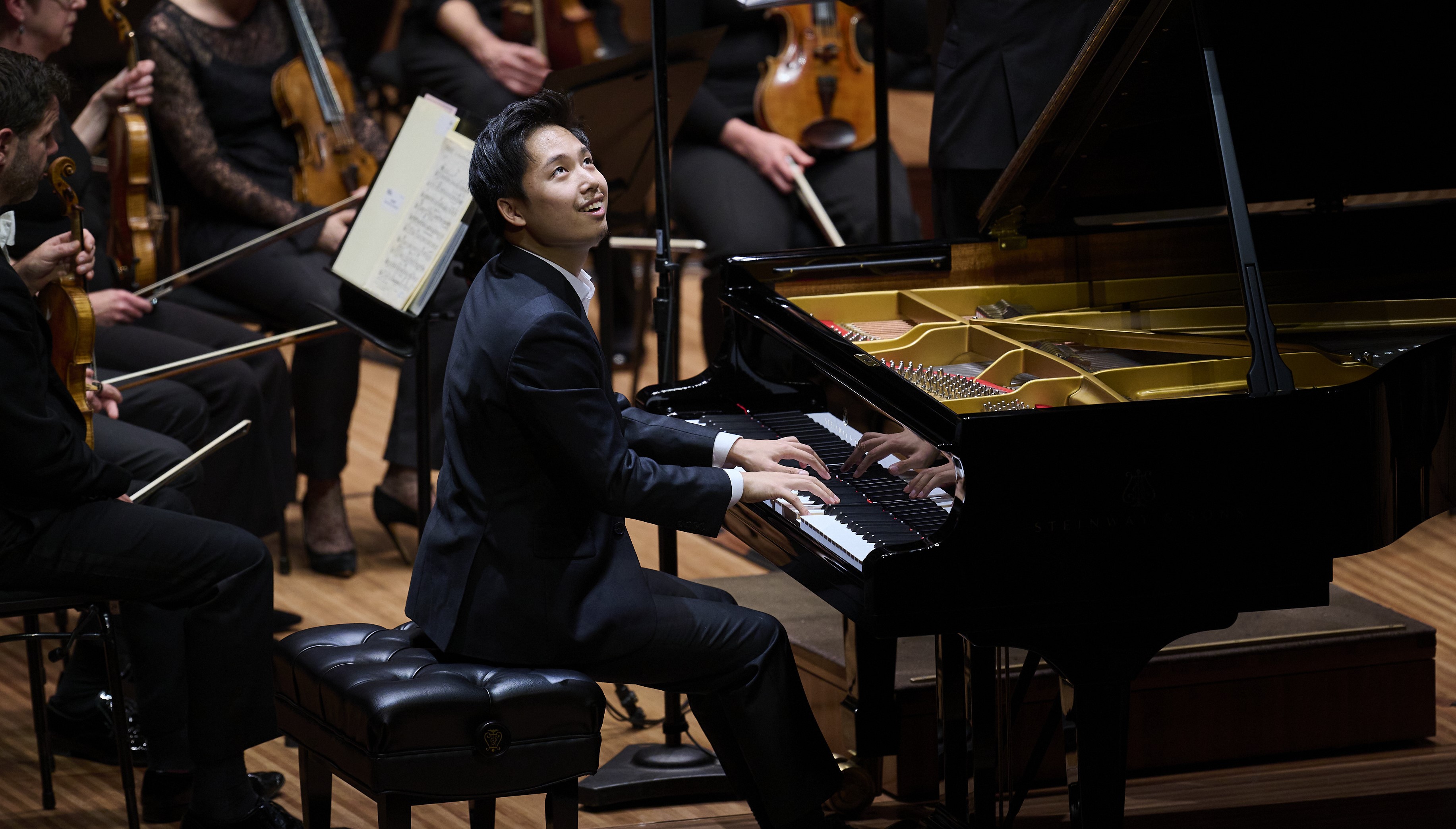 A Korean man wearing a black suit sits at a grand piano looking up and smiling