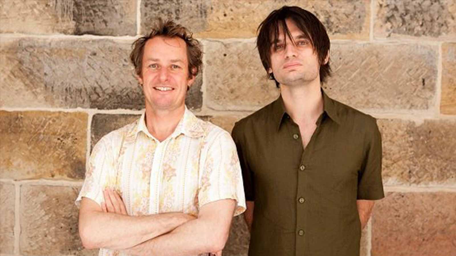 Two men in short-sleeved shirts standing outside a brick wall.