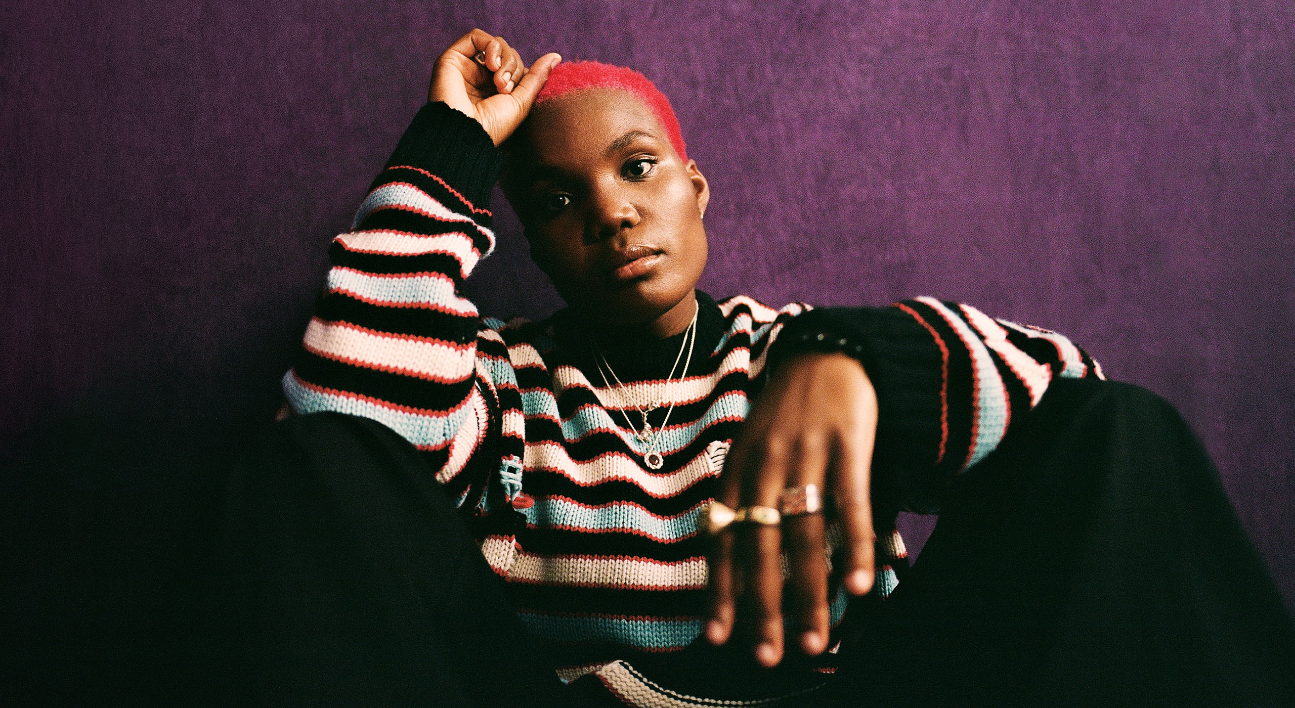 Arlo Parks wearing a striped knitted sweater, sitting on the ground against a dark purple wall.