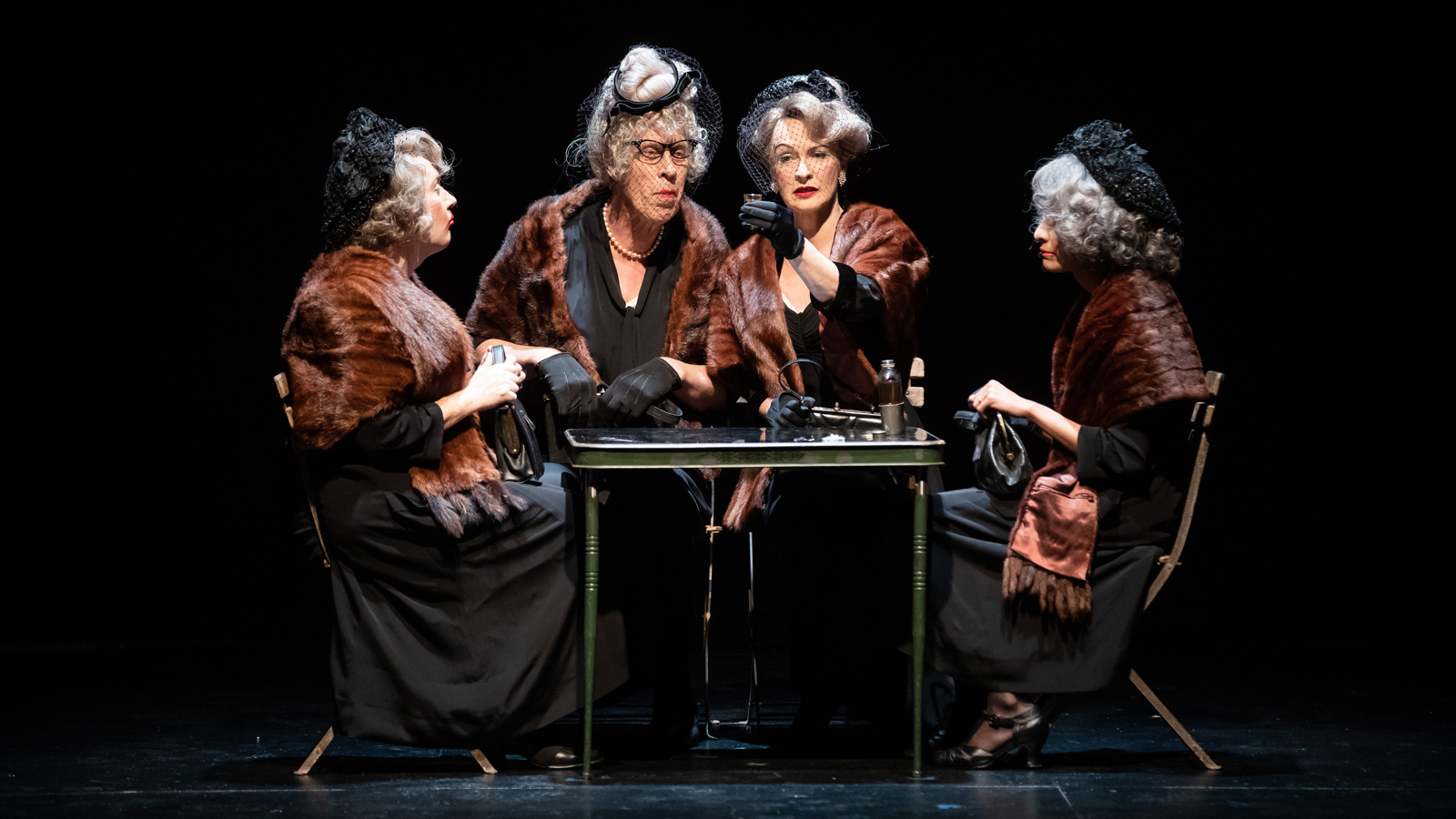 Two men dressed as a woman with two women sitting around a table and having a discussion.