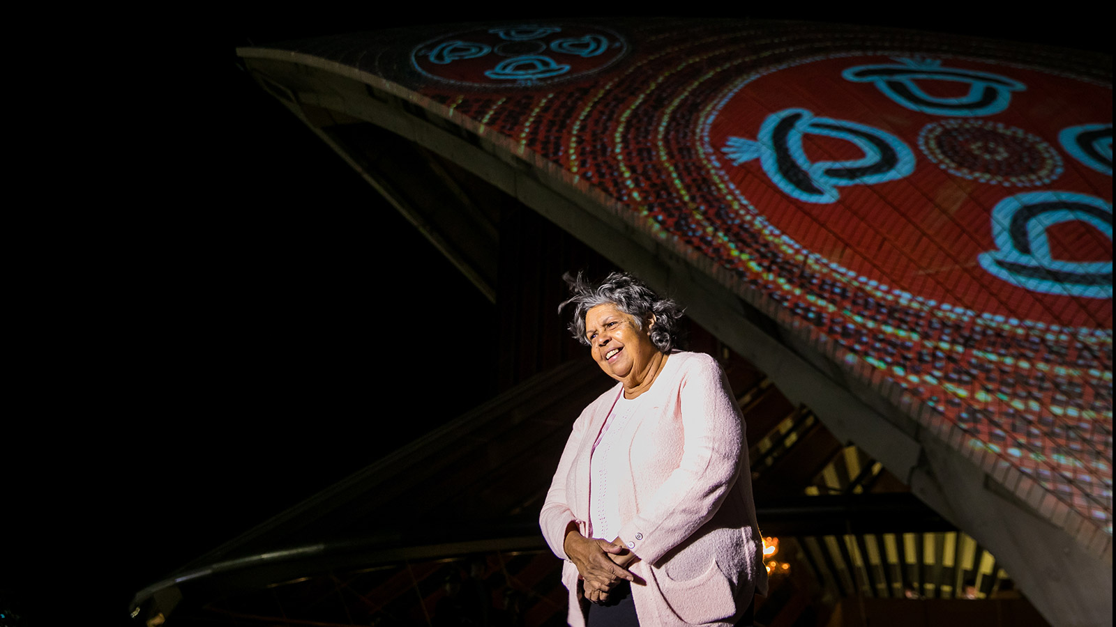 A woman in a white jacket against a textured colorful visual projected onto the Sydney Opera House's sails, smiling.