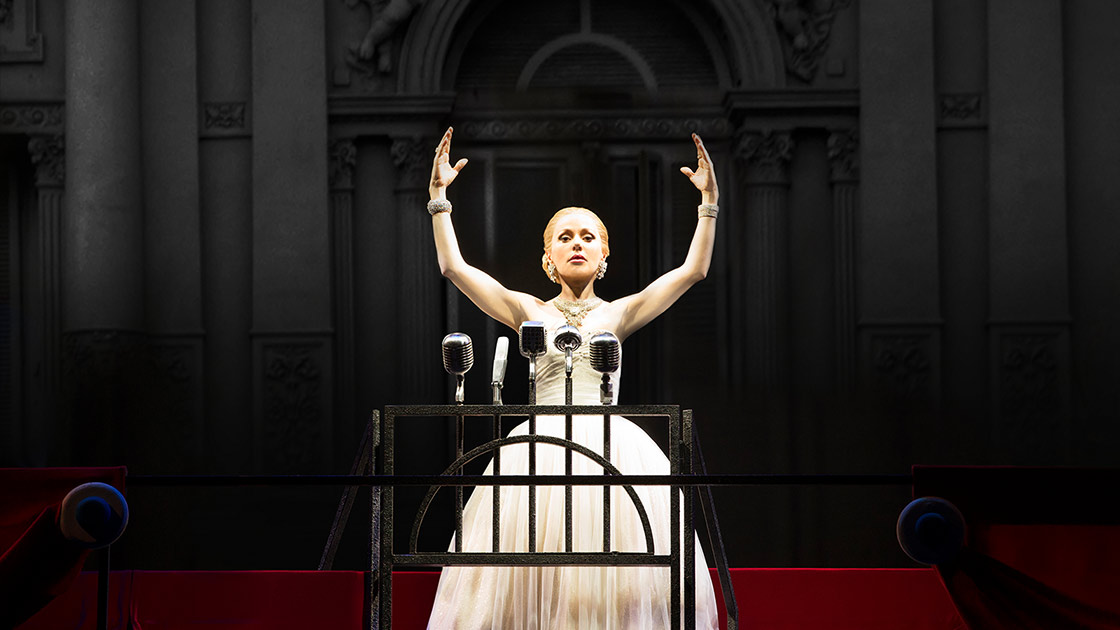 An actress holding her arms up, dressed in a white dress and stood behind a podium with microphones.