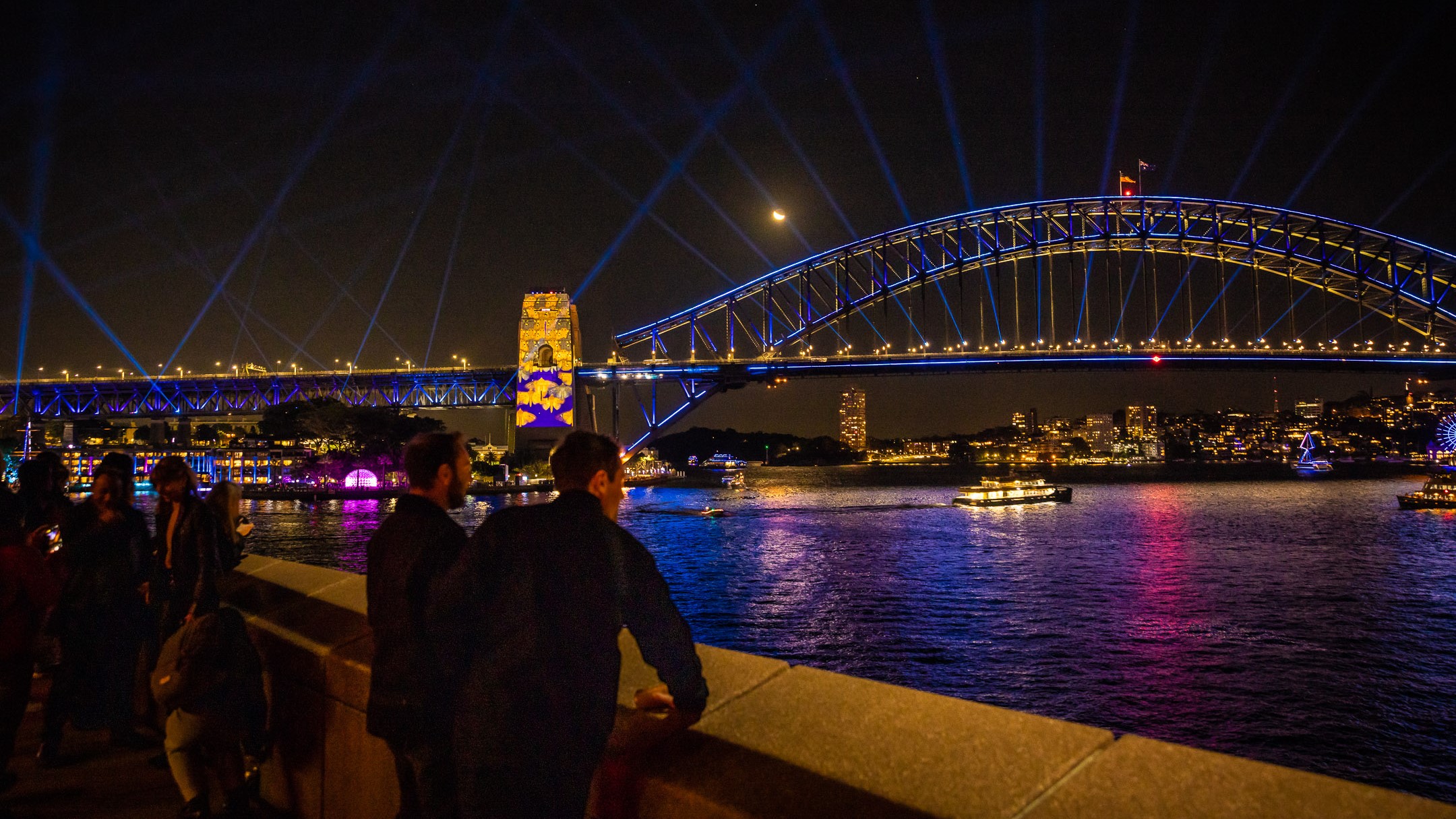 Two white men look out over Sydney Harbour with the Sydney Harbour Bridge lit up in blue in front of them.