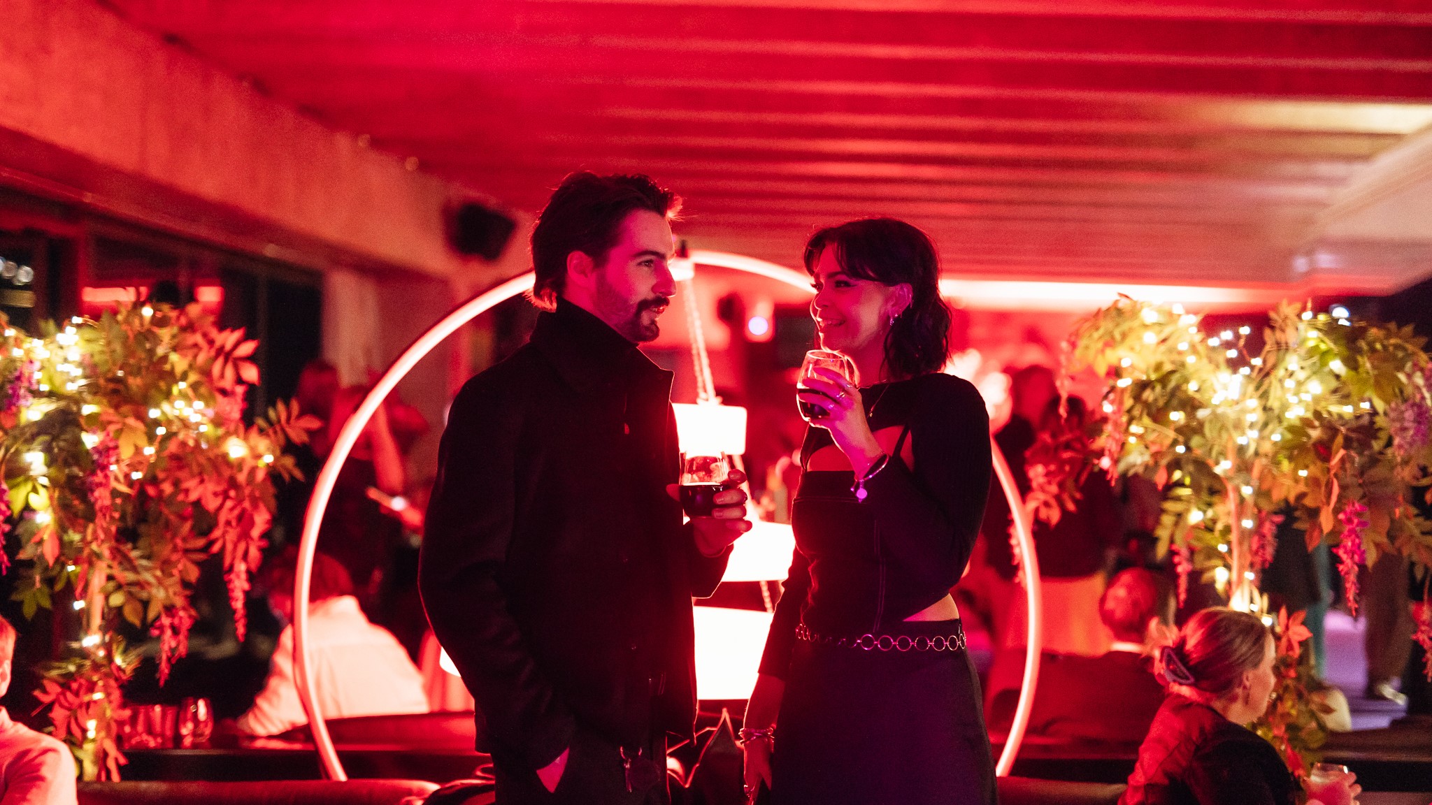 A white man with brown hair and a beard stands next to a white woman with short brown hair talking and drinking red wine, surrounded by red ambient light.