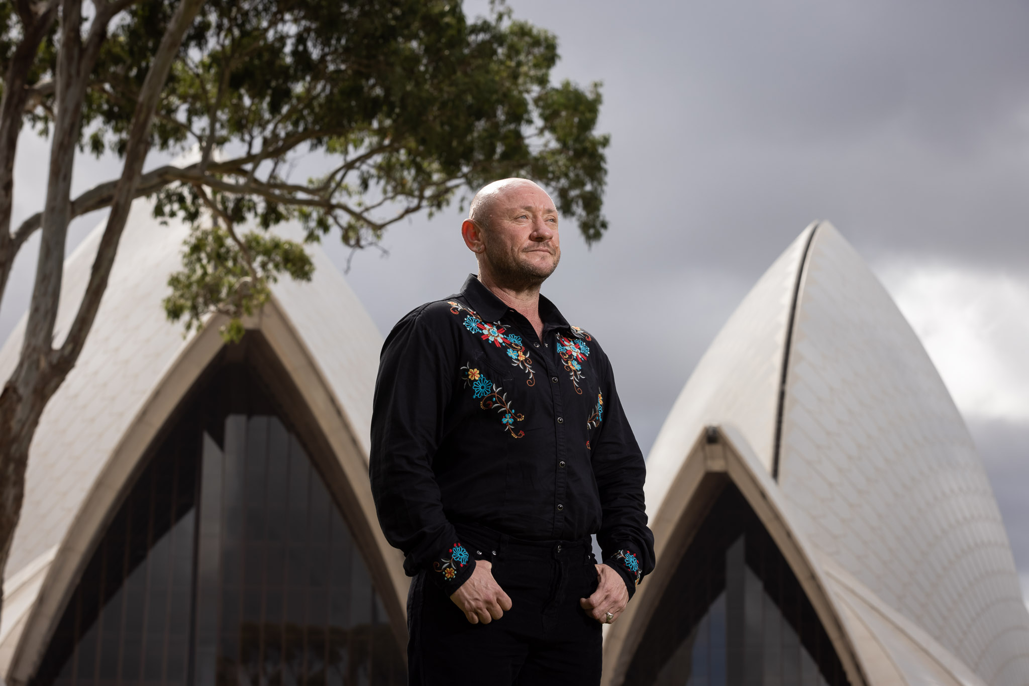 A man wearing a black embroidered shirt standing outside Sydney Opera House.