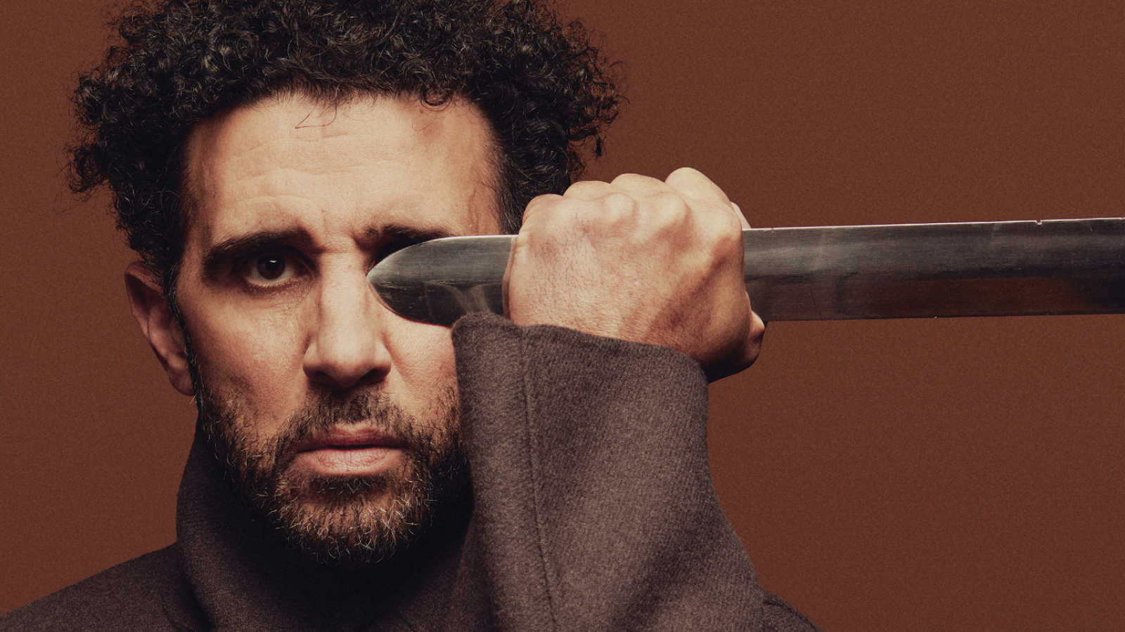 A man with stubble beard and curly hairs holding a sword blade.