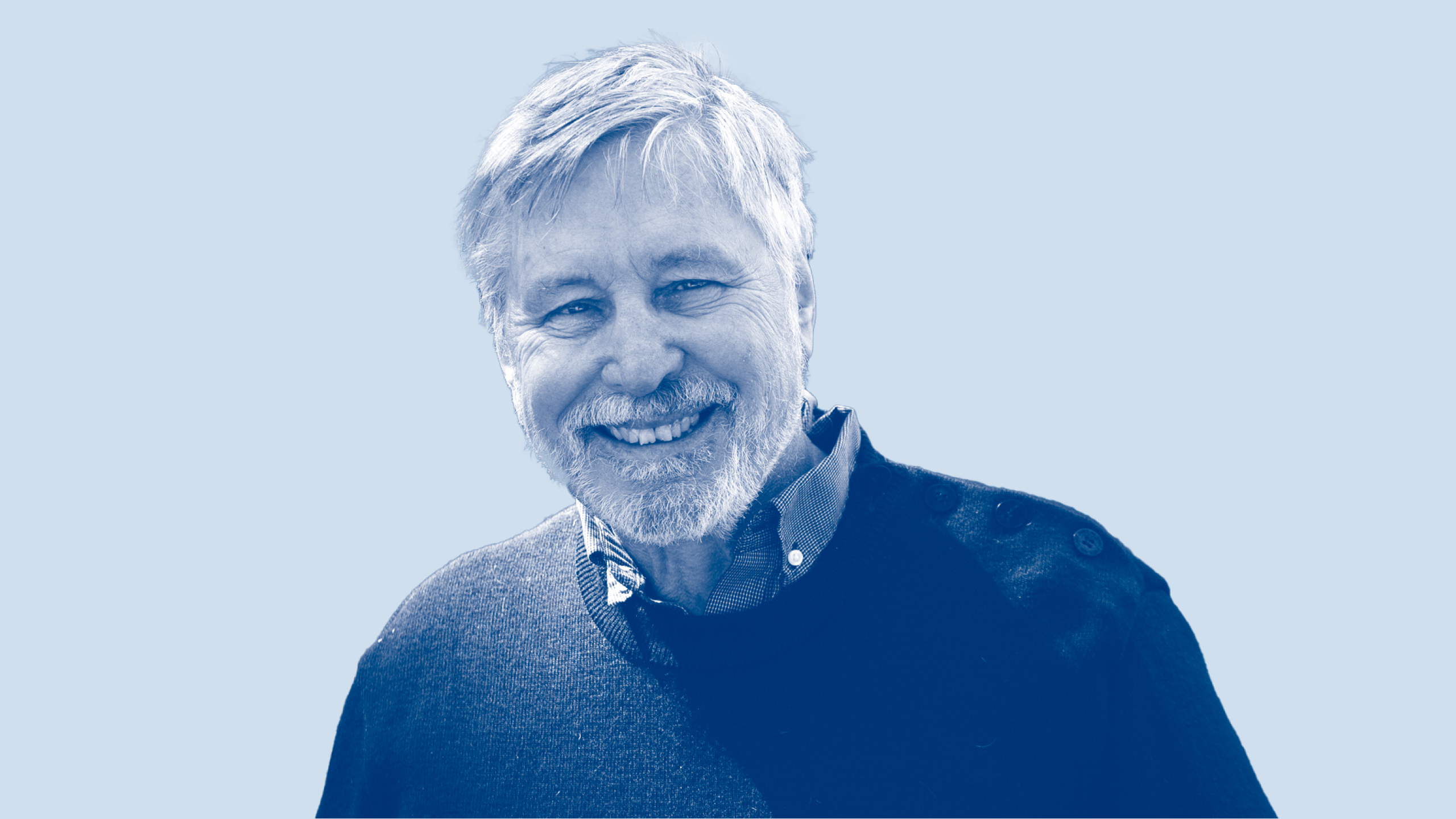A white man with white hair and a beard smiling. He wears a blue sweater.