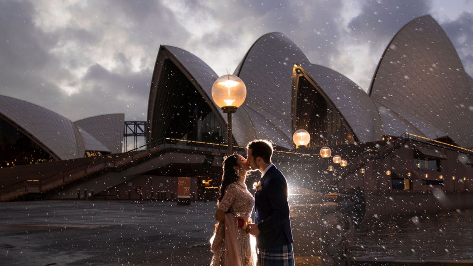 A woman in a wedding dress and a man in a suit kissing outside of the Sydney Opera House.