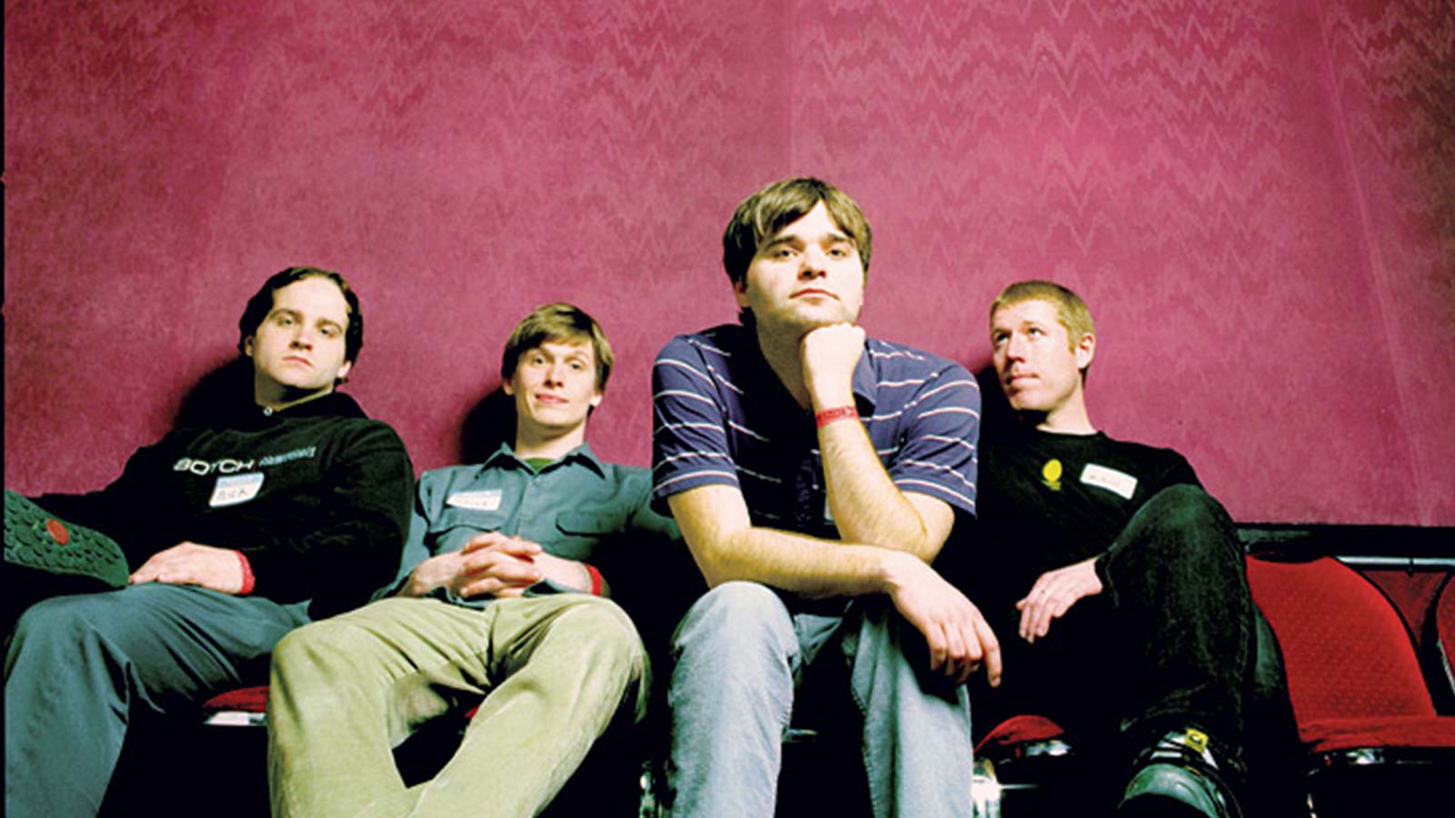 A group of four men seated on red chairs in front of a pink background.