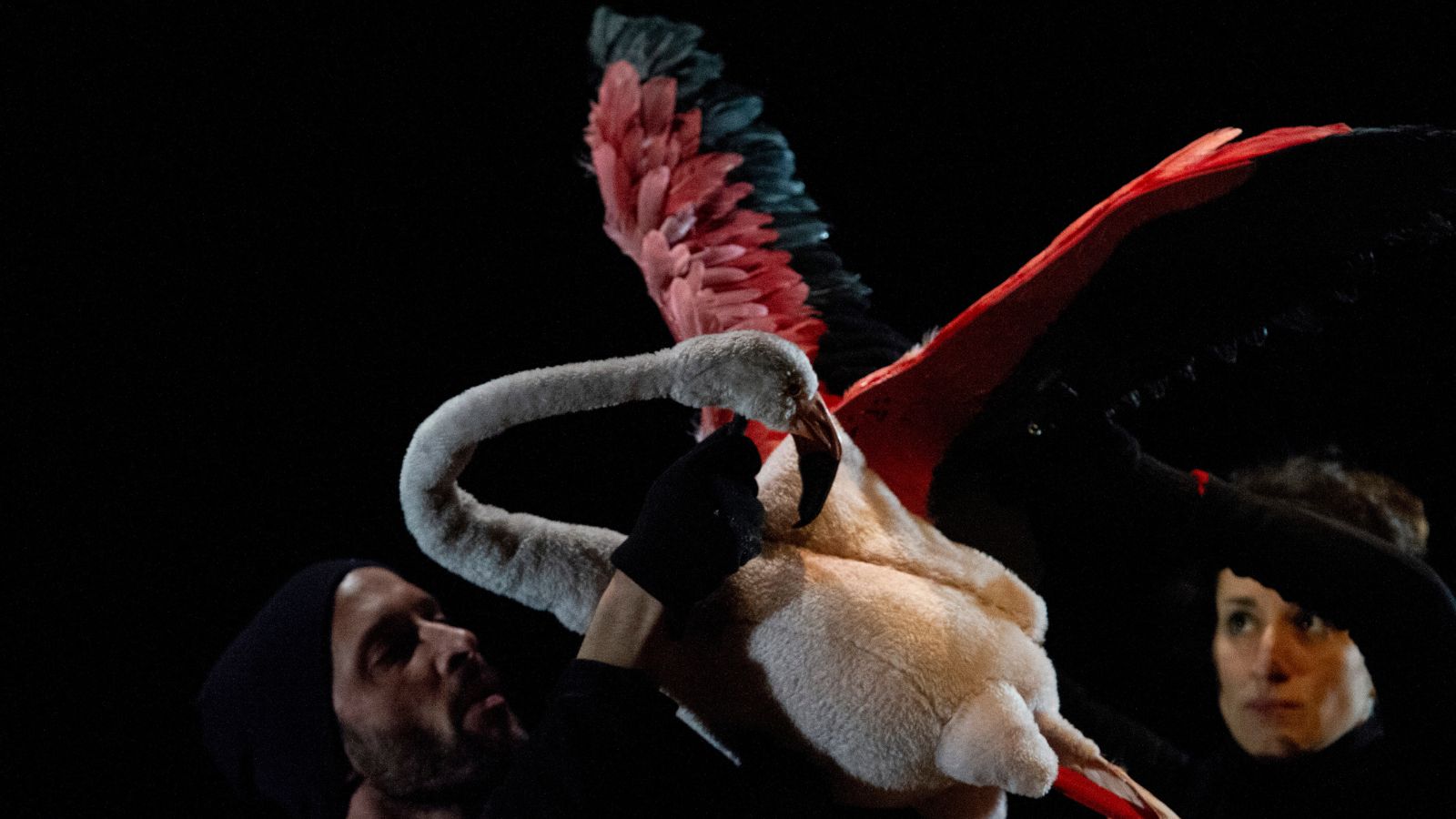 A puppet of a pink flamingo flies through the air controlled by two ladies in black.