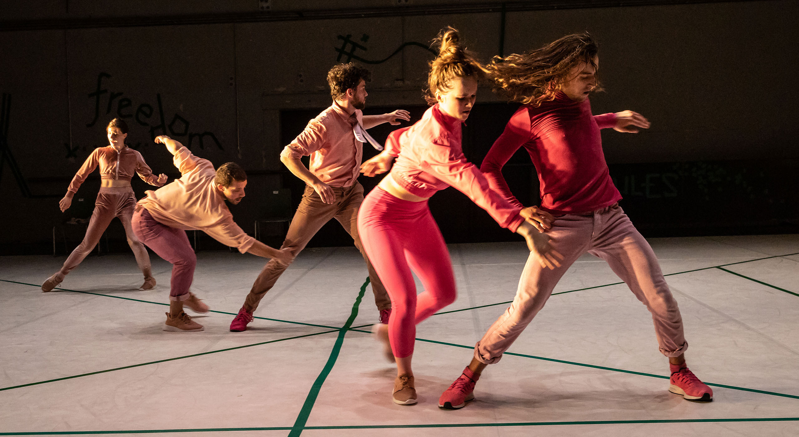 Five dancers wear pink and stand in a diagonal line caught in movement.