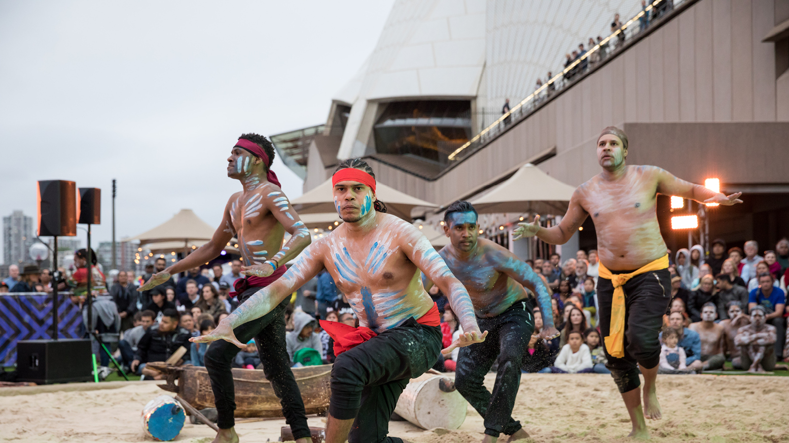 Men in sand pit performing indigenous dance in the homeground of Sydney opera house.