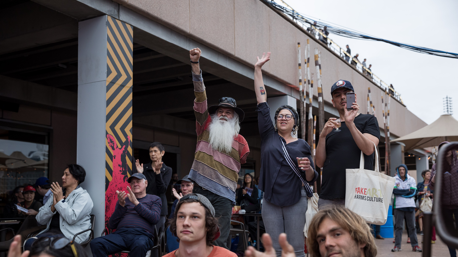 A Man with long white beard and a woman standing in the Homeground with their hands up.