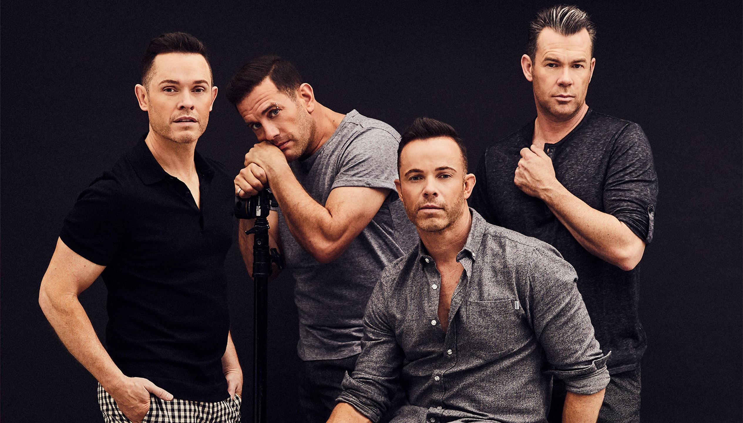 Four white men wearing various shades of black, white and grey are posed in front of a black background. Three men are standing and one is seated on a stool.