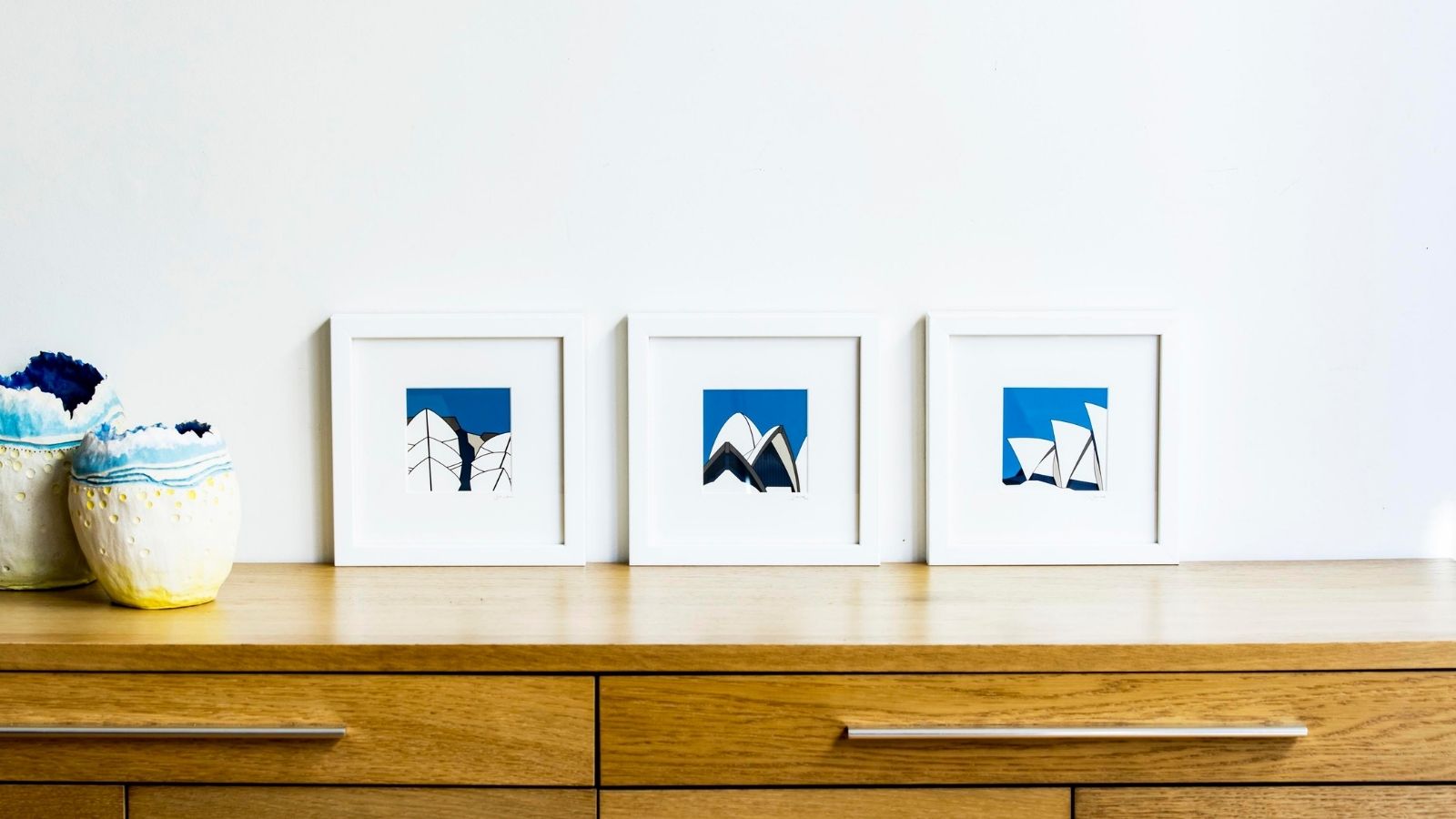 Three framed blue pictures resting on a wooden cabinet against a white wall.