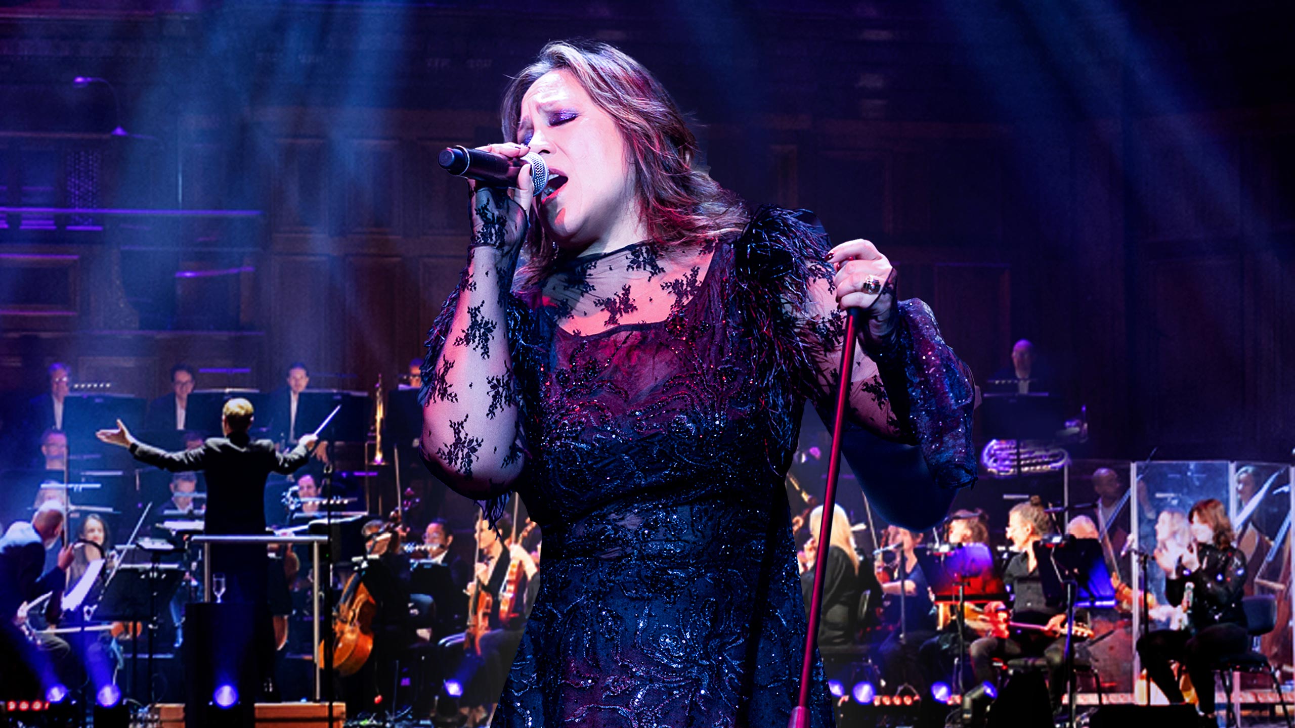 A Filipino-Australian woman with brown layered, shoulder-length hair holds a microphone in her right hand and the microphone stand in her left hand. She's singing on stage in front of an orchestra.