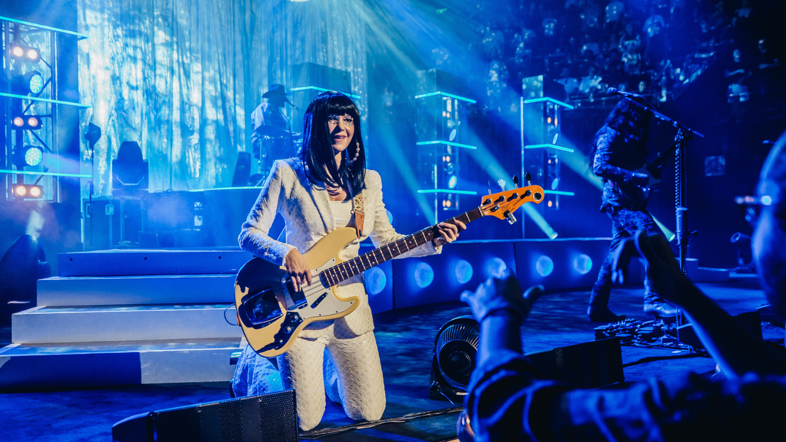 Woman with long dark hair knees while playing bass guitar on stage