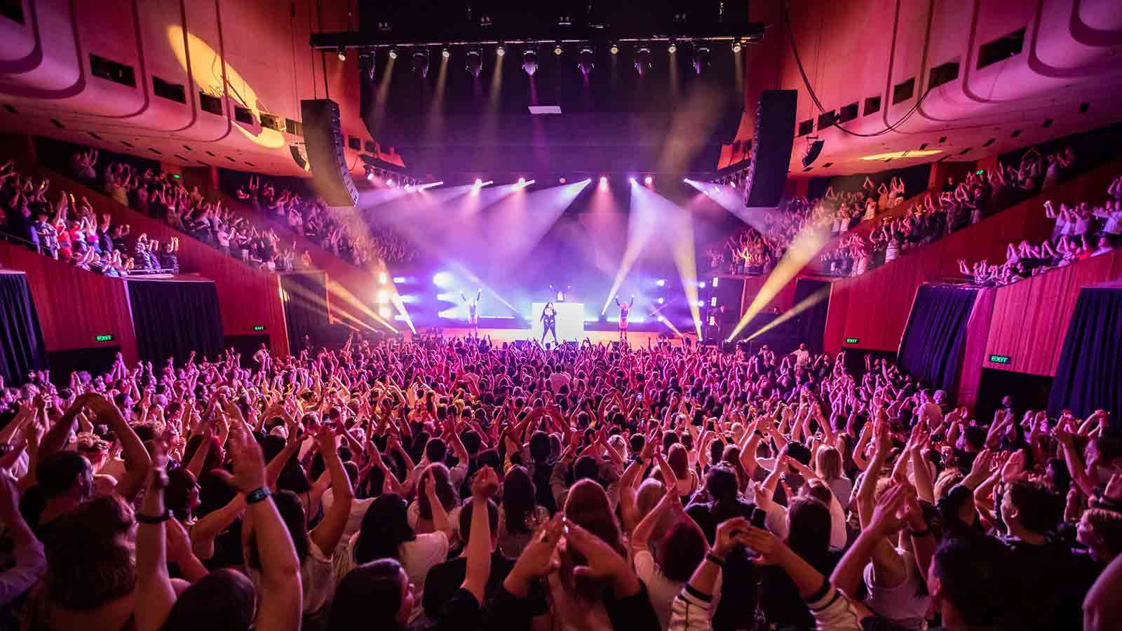 A lively audience at an inside venue, lit by pink strobes.