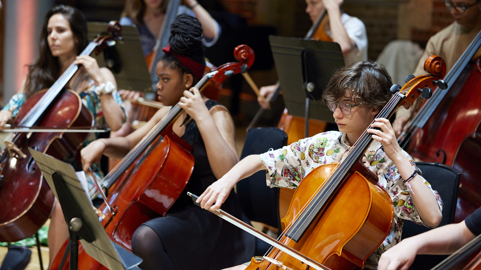 Two rows of young people seated in a room, playing cellos.