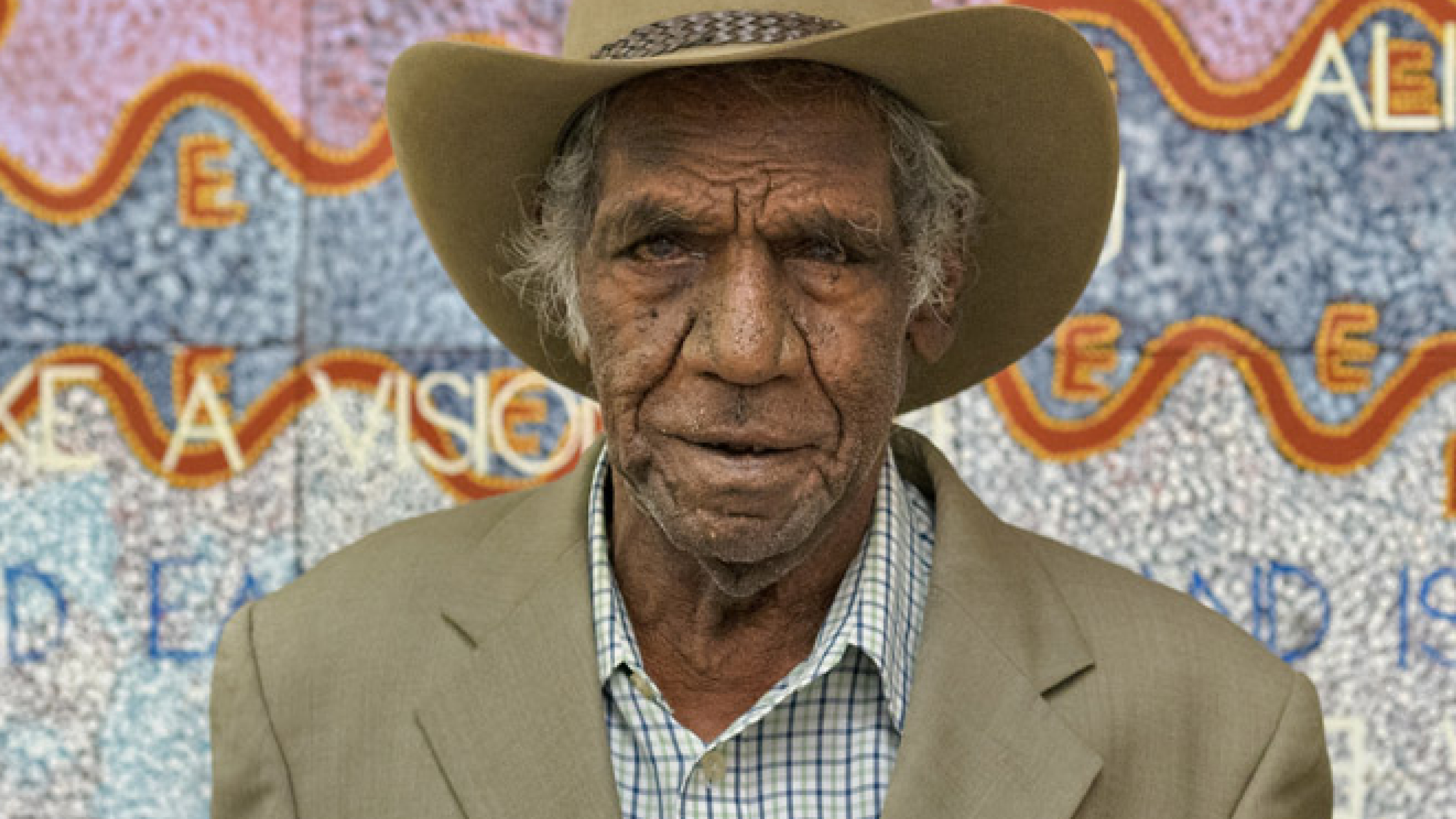 Michael Nelson Jagamara wearing a beige suit jacket over a blue and white checked shirt and wide brim hat, standing in front of an artwork in pastel colours.