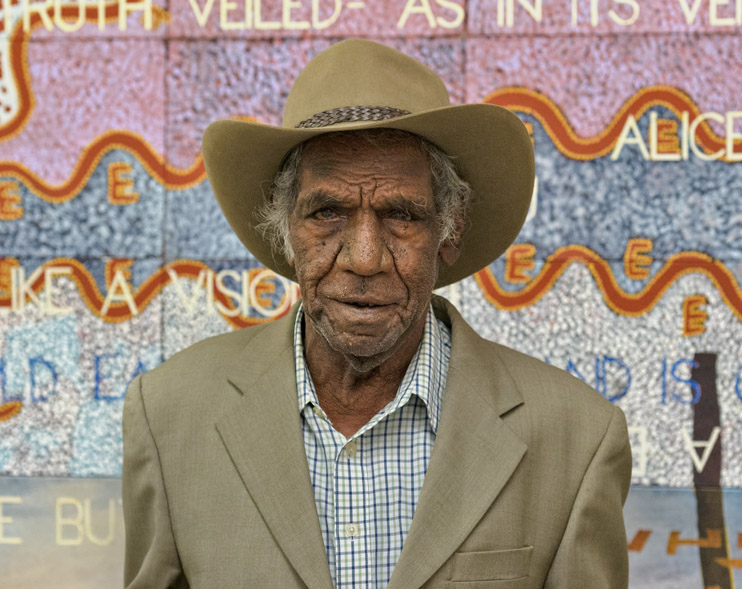 Michael Nelson Jagamara wearing a beige suit jacket over a blue and white checked shirt and wide brim hat, standing in front of an artwork in pastel colours.