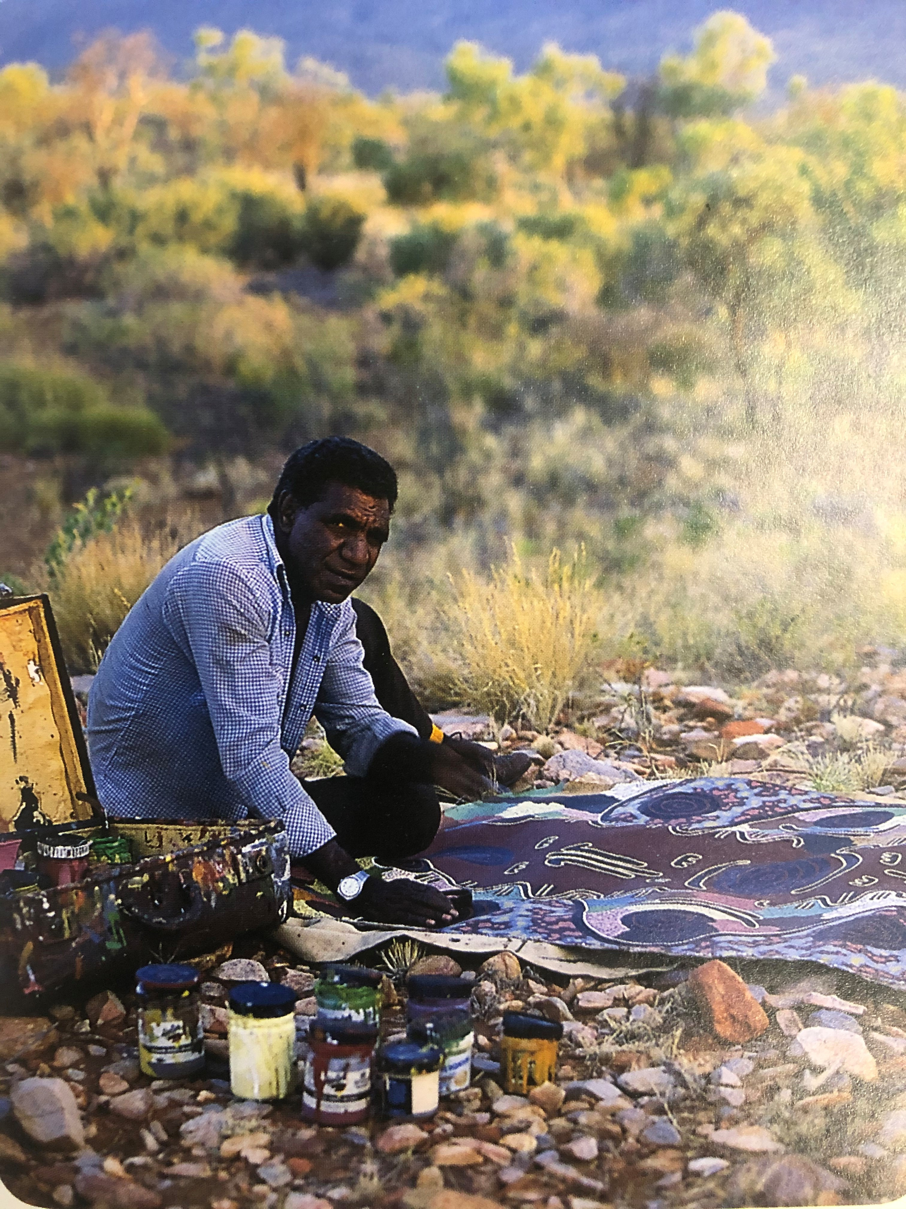 Michael Nelson Jagamara sitting on the ground, surrounded by bushland, leaning over a painting, laid out in front of him.